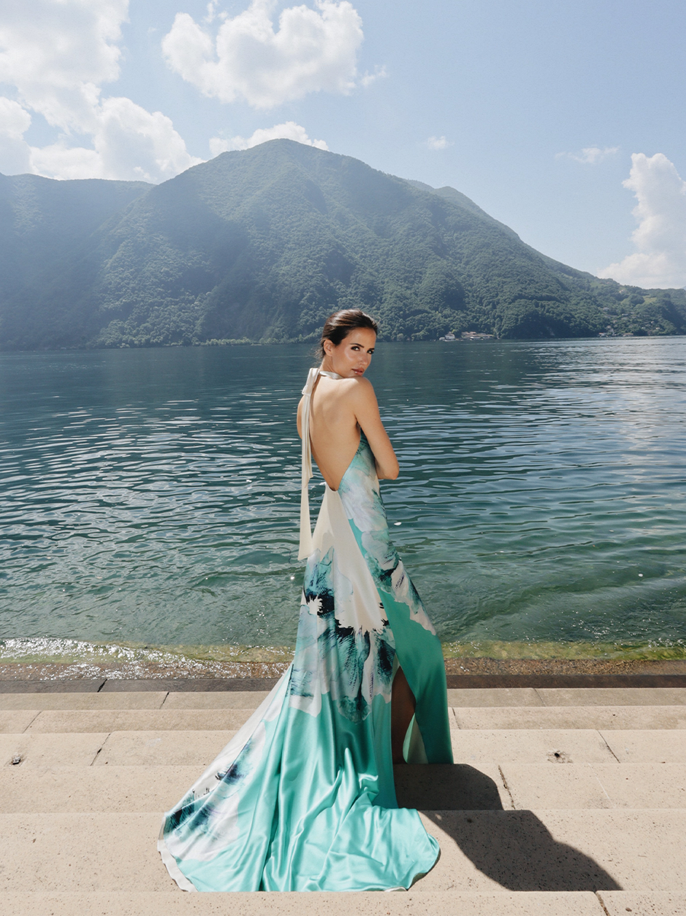Una mujer con un vestido suelto sin espalda se encuentra de pie sobre unos escalones de piedra junto a un lago, con montañas y un cielo parcialmente nublado de fondo. El vestido presenta tonos verdes y azules con un estampado floral.