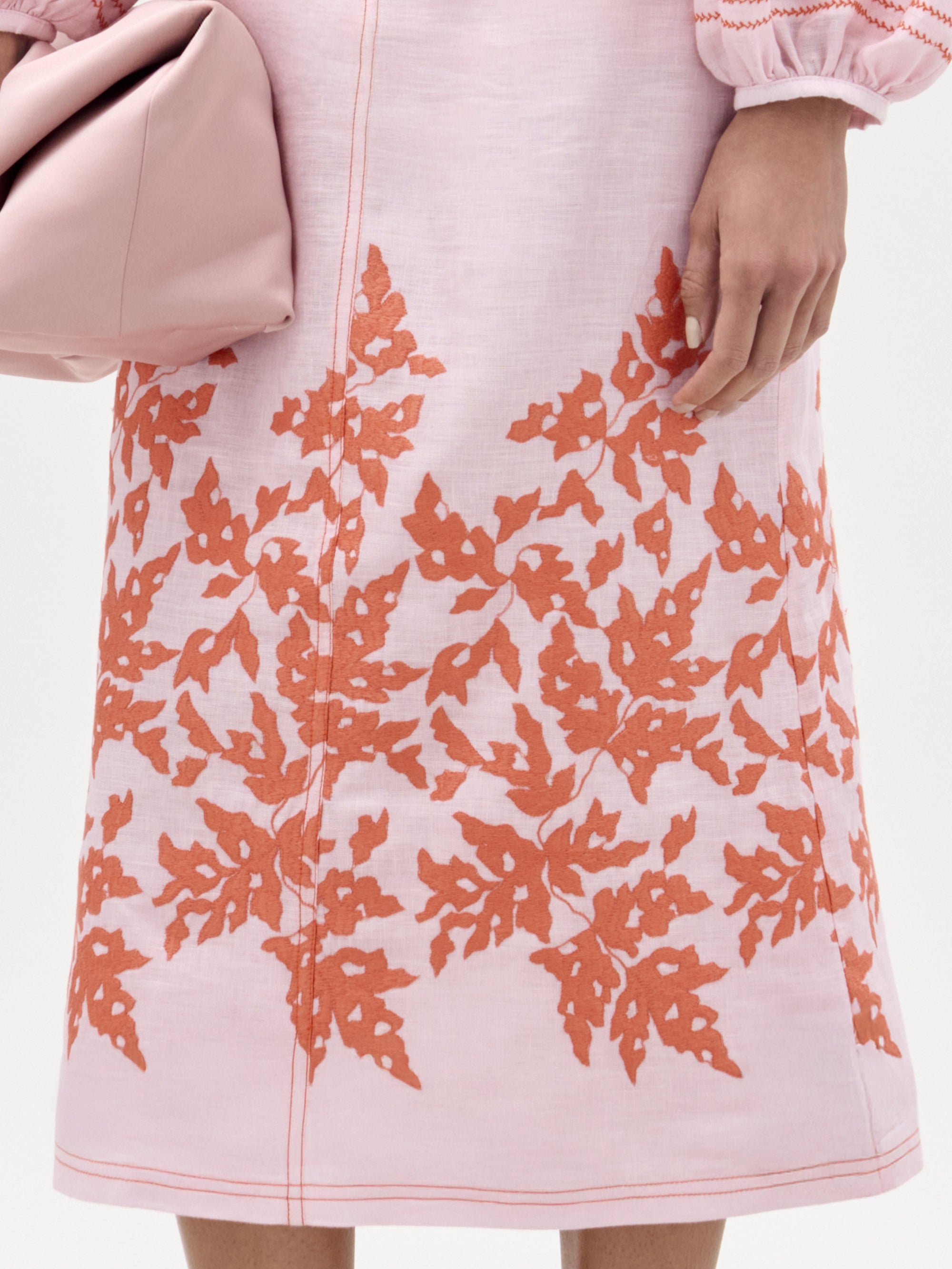 Una persona que lleva la falda Layan -de color rosa claro con un bordado de hojas naranjas cerca del dobladillo- sostiene una bolsa de color rosa suave. Solo se ven la parte inferior del torso y la mano.