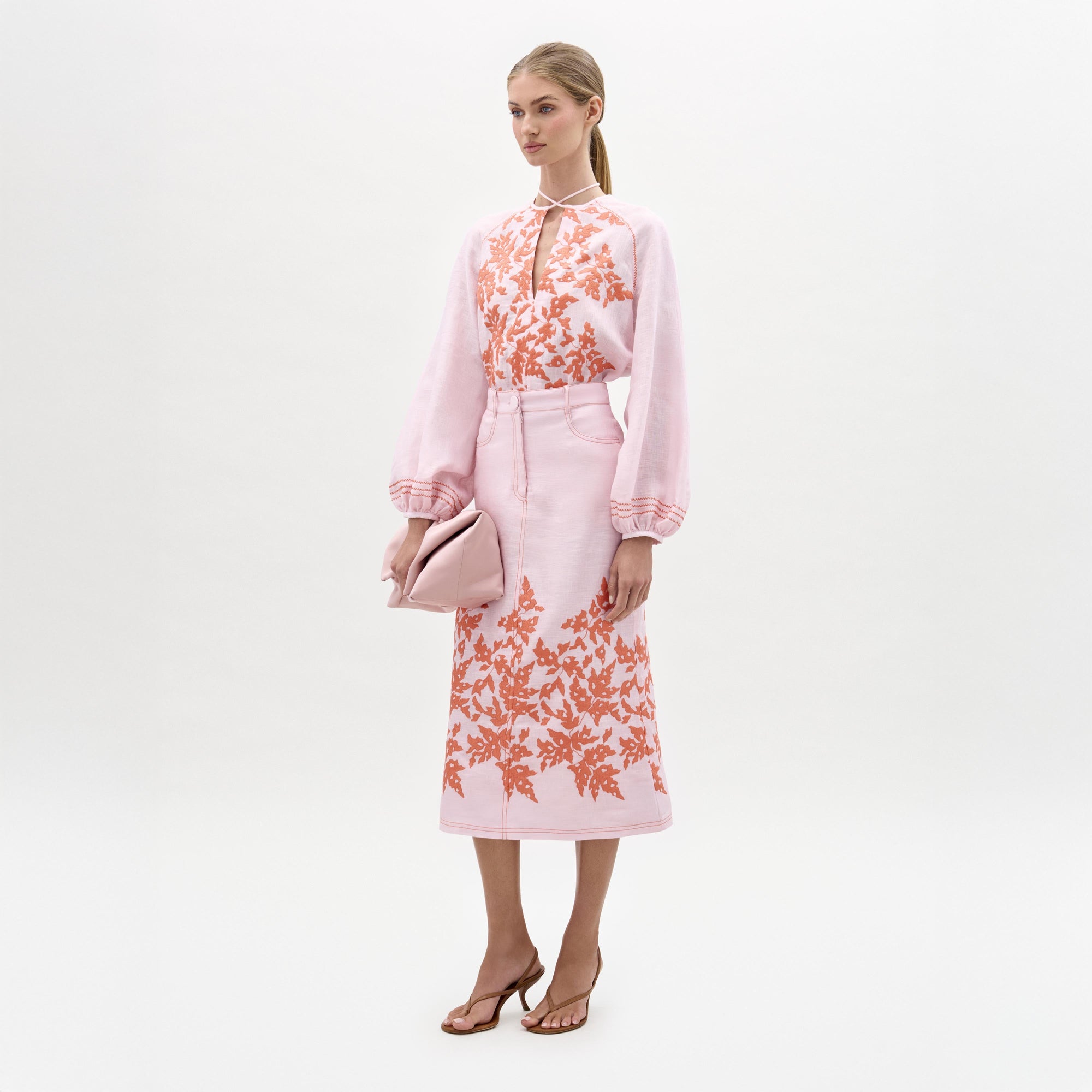 Una mujer, sobre fondo blanco, lleva una blusa rosa claro con motivos florales naranjas, la falda Layan a juego, sandalias de tacón y un bolso de mano rosa doblado.