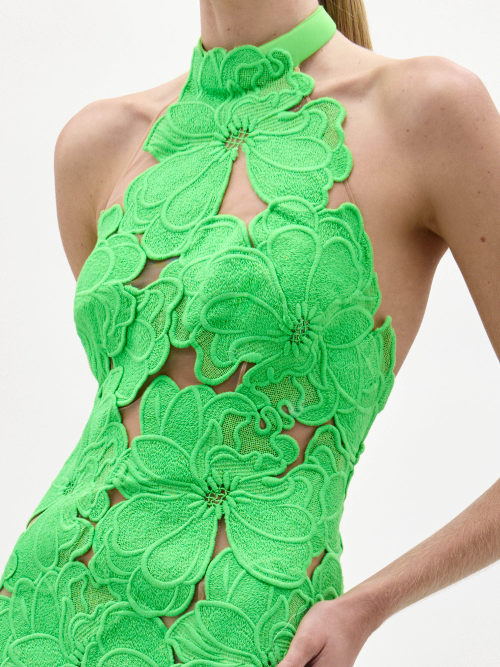 Una persona lleva el vestido Mab, un modelo sin mangas y cuello halter de encaje floral verde brillante con llamativos motivos florales y recortes transparentes, sobre un fondo blanco liso.