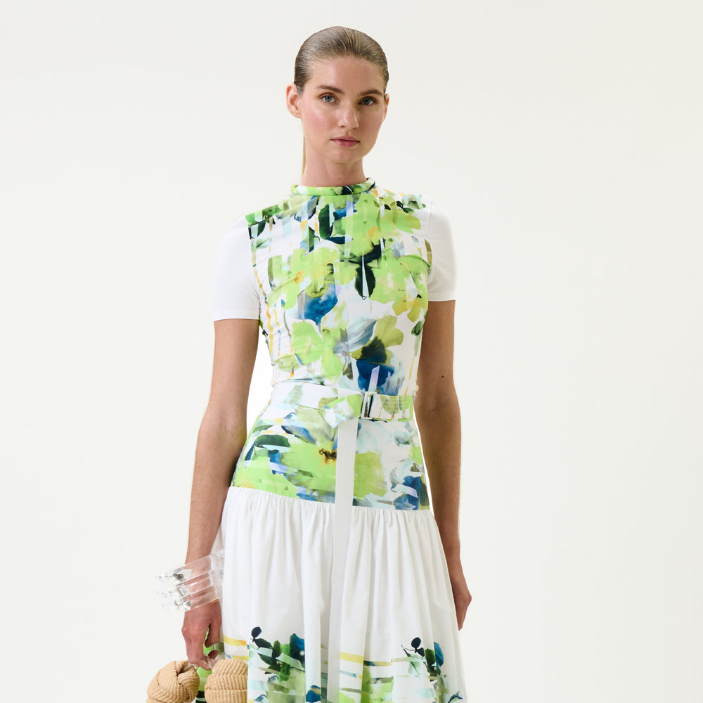 A model wearing a sleeveless white dress with a vibrant green, blue, and white abstract floral print on the bodice and skirt, styled with a white t-shirt underneath and holding a straw hat.