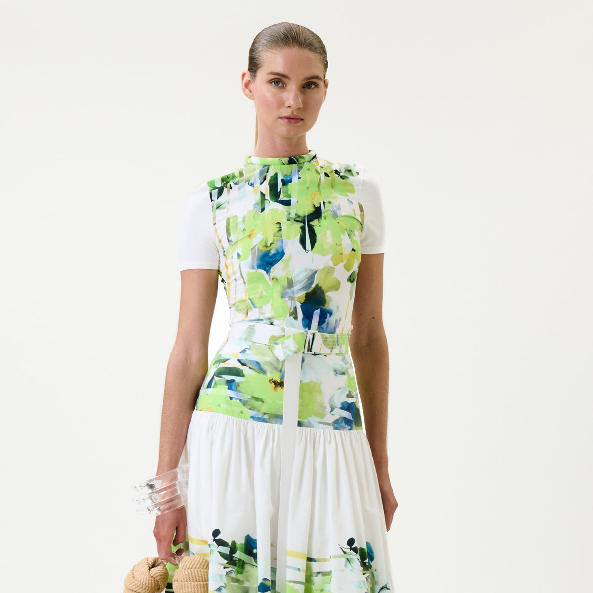 A model wearing a sleeveless white dress with a vibrant green, blue, and white abstract floral print on the bodice and skirt, styled with a white t-shirt underneath and holding a straw hat.