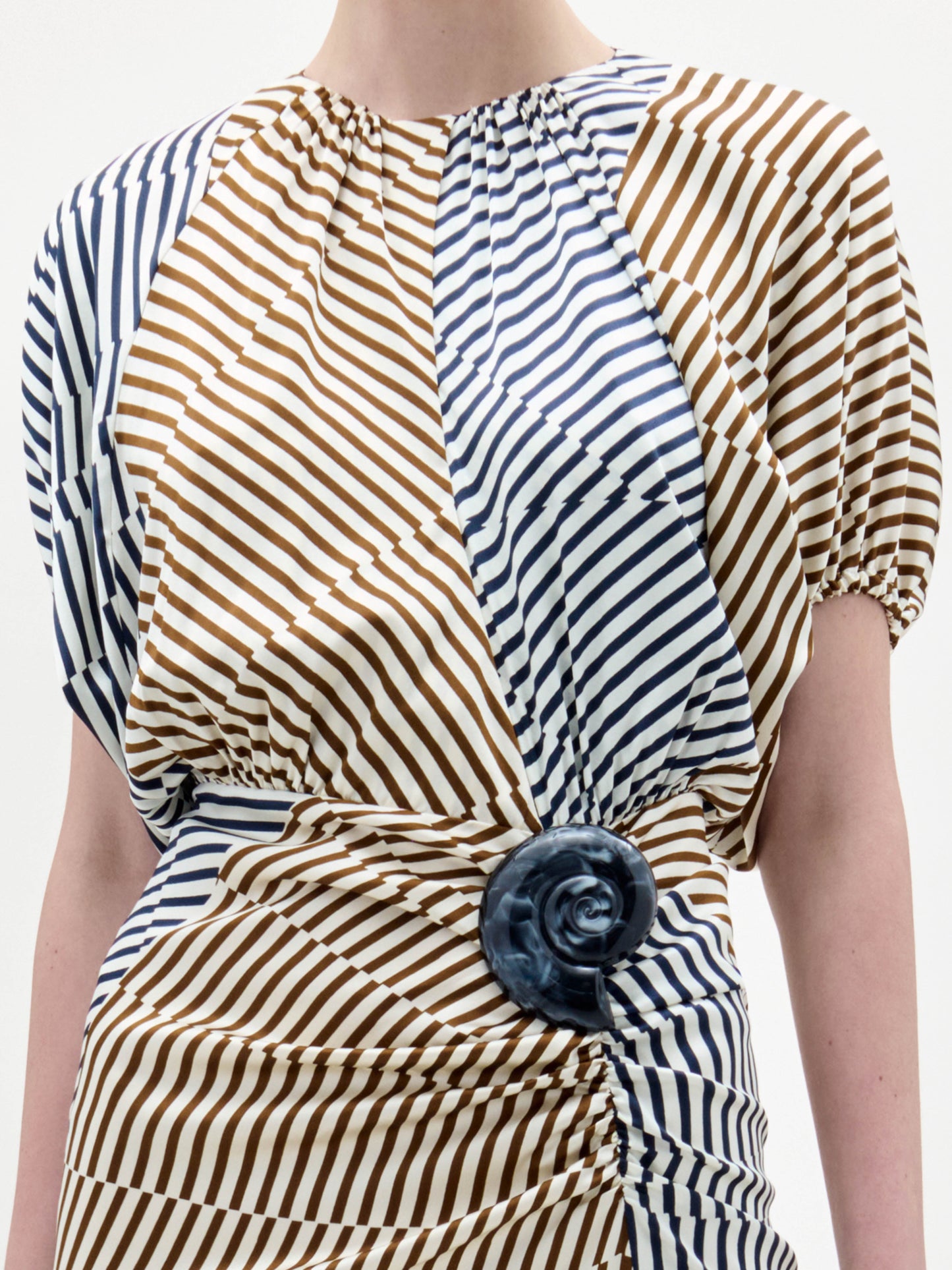 Una persona lleva el vestido Kala, de manga corta, con rayas diagonales marrones y azul marino, tejido fruncido y un gran acento circular azul oscuro en la cintura. El fondo es blanco liso.