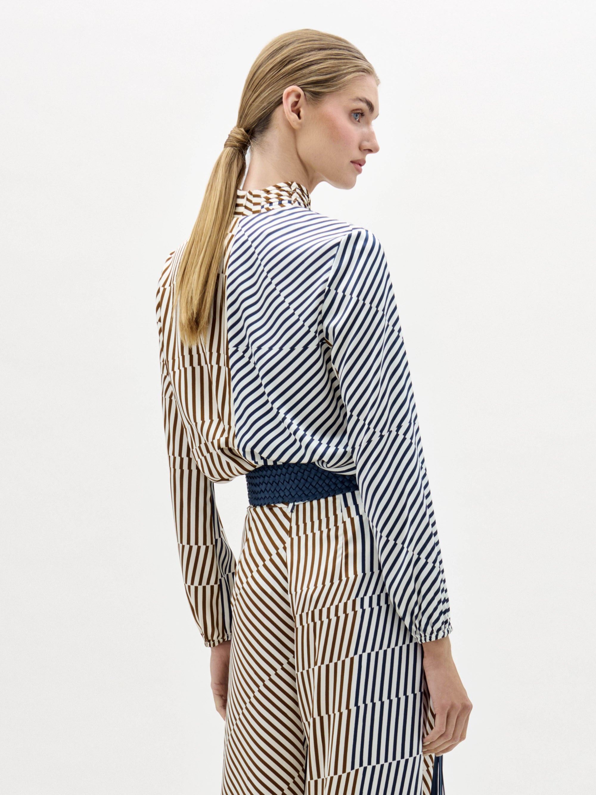 Una mujer con el pelo rubio liso recogido en una coleta baja, de pie y de lado, lleva la blusa Jasper a rayas verticales y diagonales en marrón, blanco y azul sobre un fondo blanco liso.