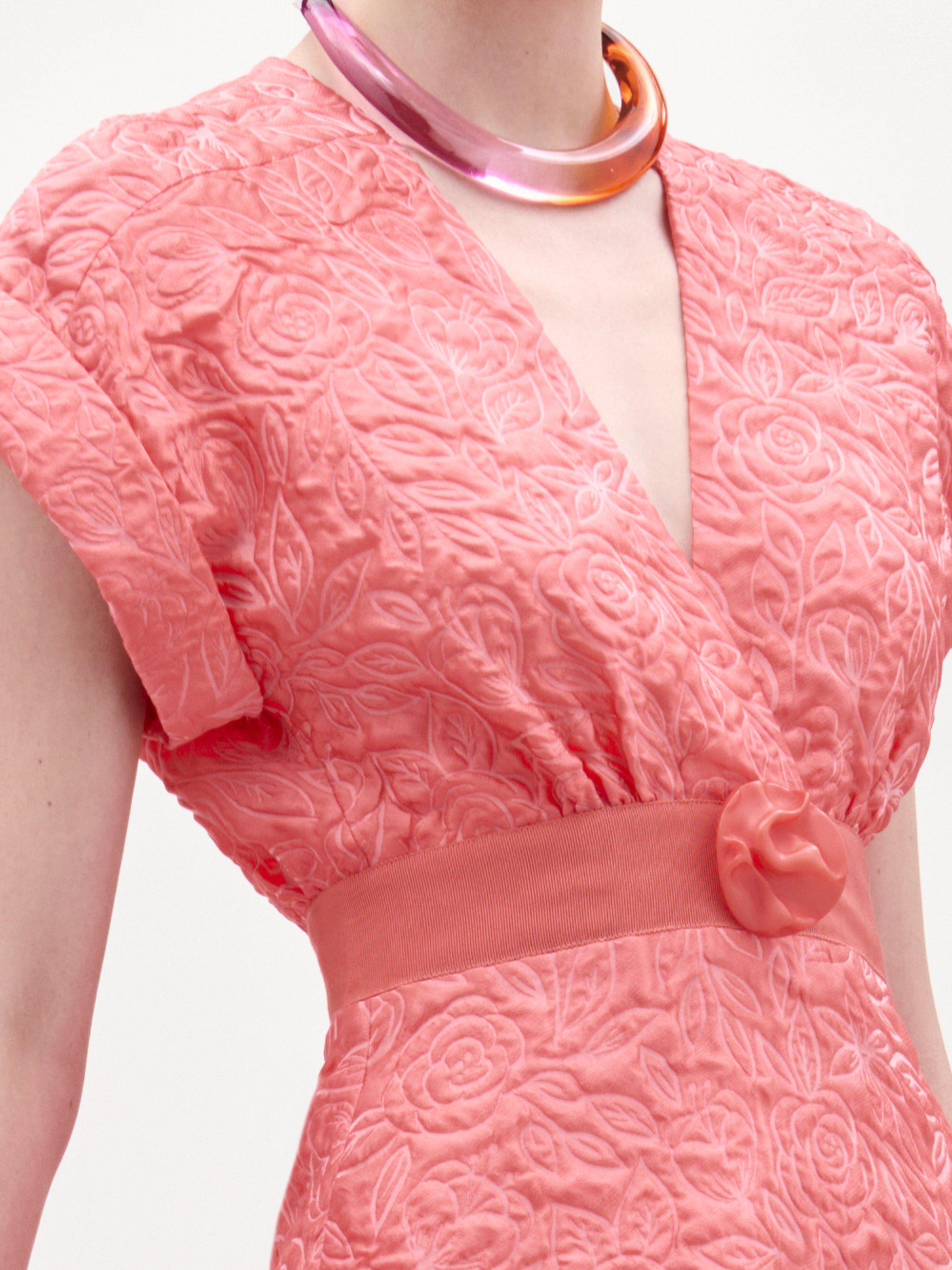 Una persona luce el vestido Metaponto -un vestido midi coral texturizado con estampado floral y cinturón a juego con hebilla redonda- que acentúa la cintura, combinado con un collar rosa brillante y grueso. Imagen recortada para mostrar solo la parte superior del cuerpo.
