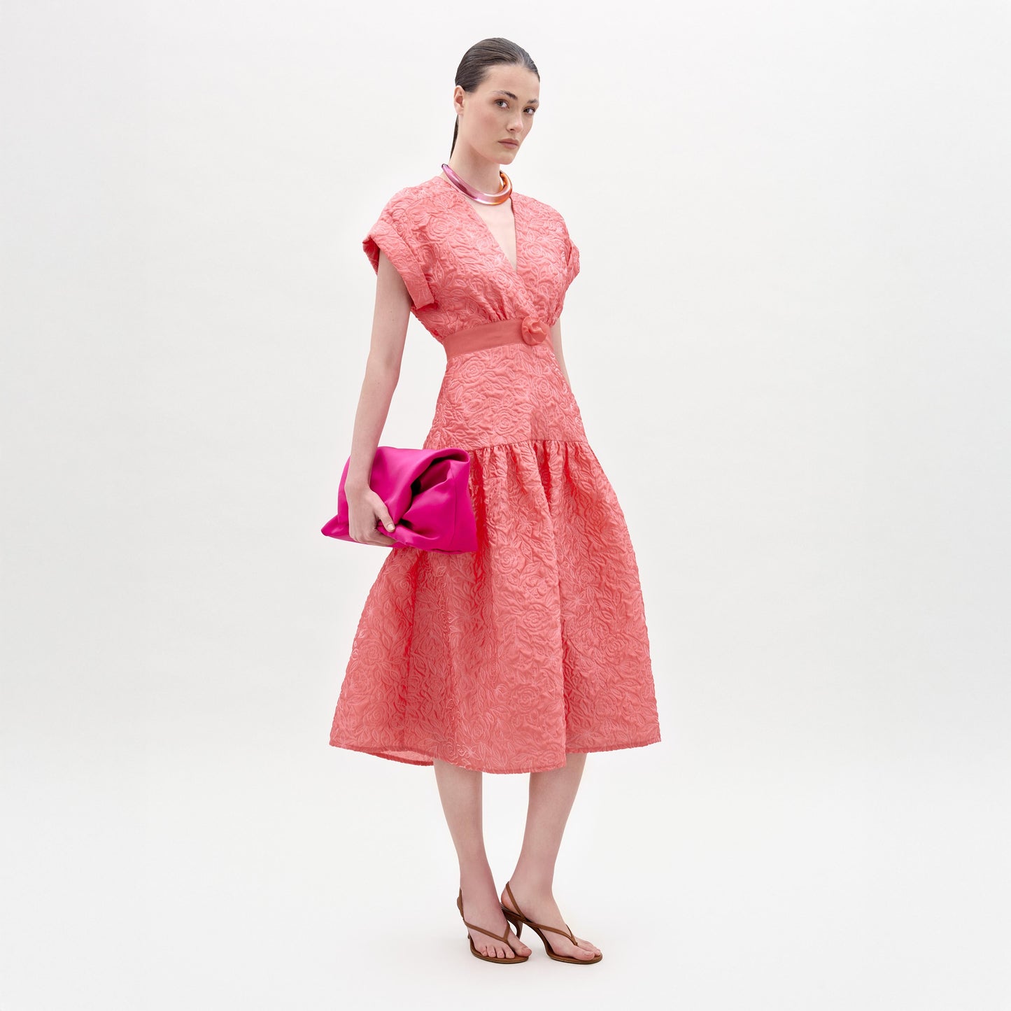 Una mujer posa sobre un fondo blanco liso con el vestido Metaponto, sandalias planas marrones, un collar rosa claro y un bolso de mano rosa brillante.