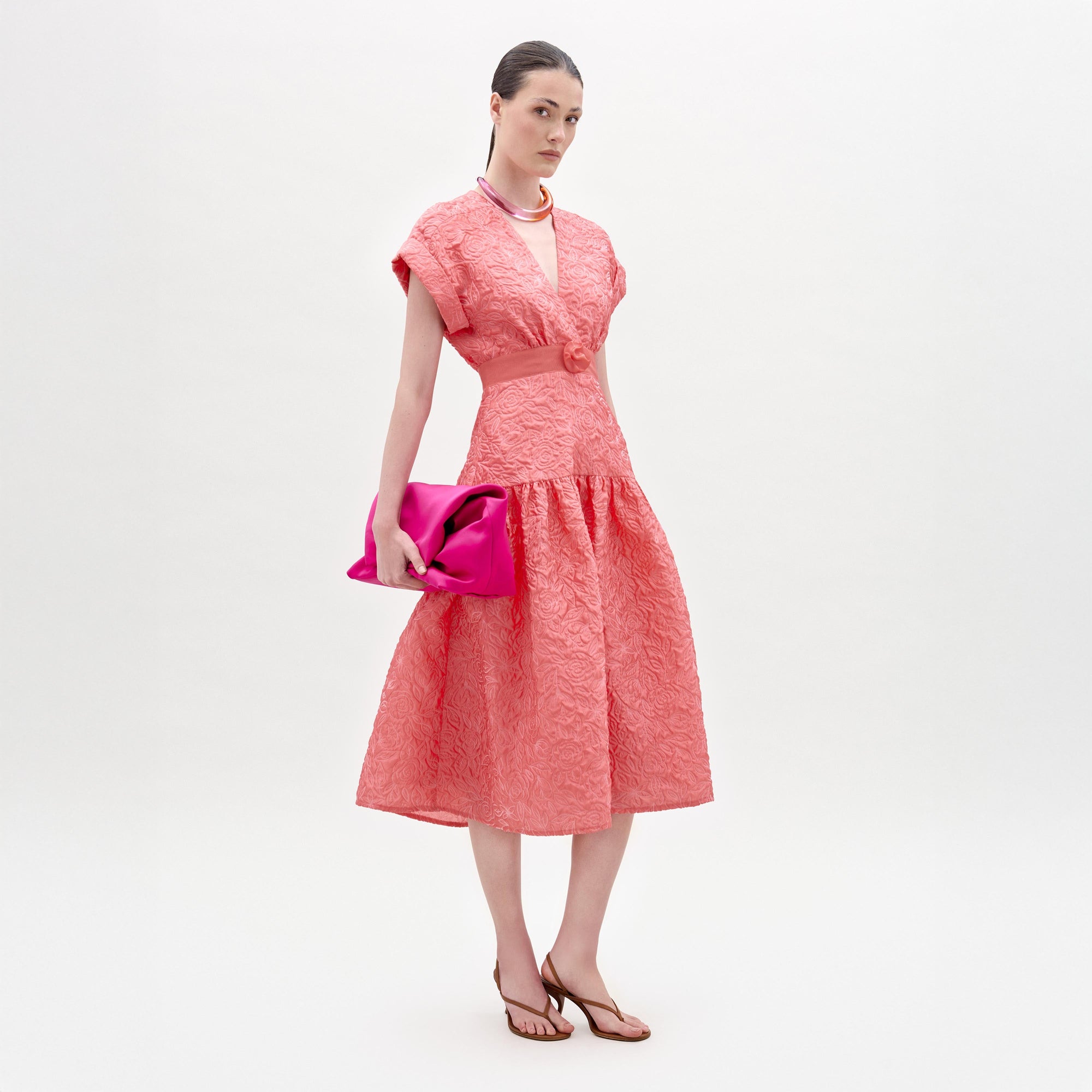 Una mujer posa sobre un fondo blanco liso con el vestido Metaponto, sandalias planas marrones, un collar rosa claro y un bolso de mano rosa brillante.
