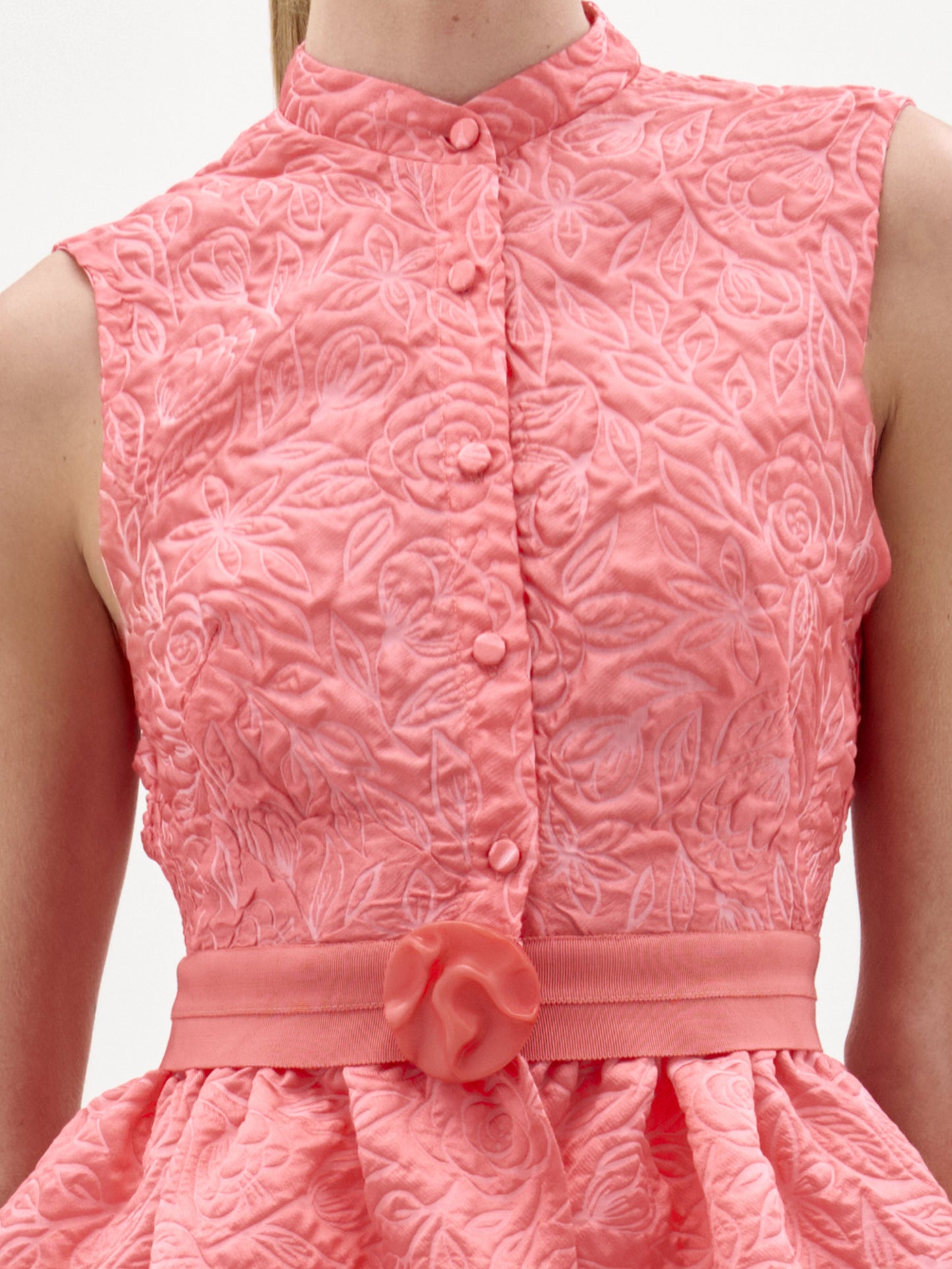Una persona lleva la blusa Colony, un top sin mangas de color rosa coral texturizado con estampado floral, botones forrados de tela en la parte delantera y un cinturón a juego con un lazo decorativo en la cintura, que recuerda a la artesanía colombiana.