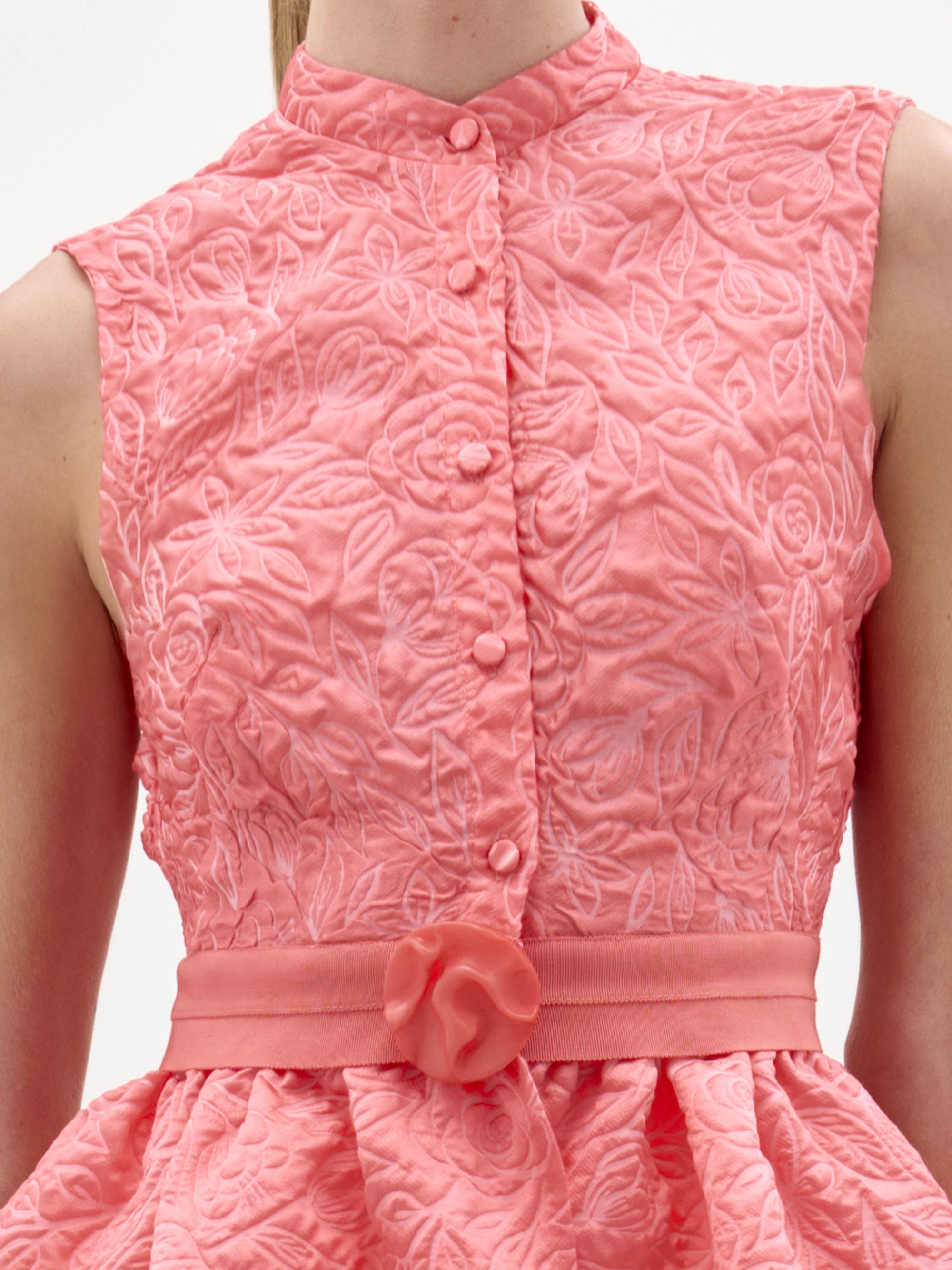 Una persona lleva la blusa Colony, un top sin mangas de color rosa coral texturizado con estampado floral, botones forrados de tela en la parte delantera y un cinturón a juego con un lazo decorativo en la cintura, que recuerda a la artesanía colombiana.