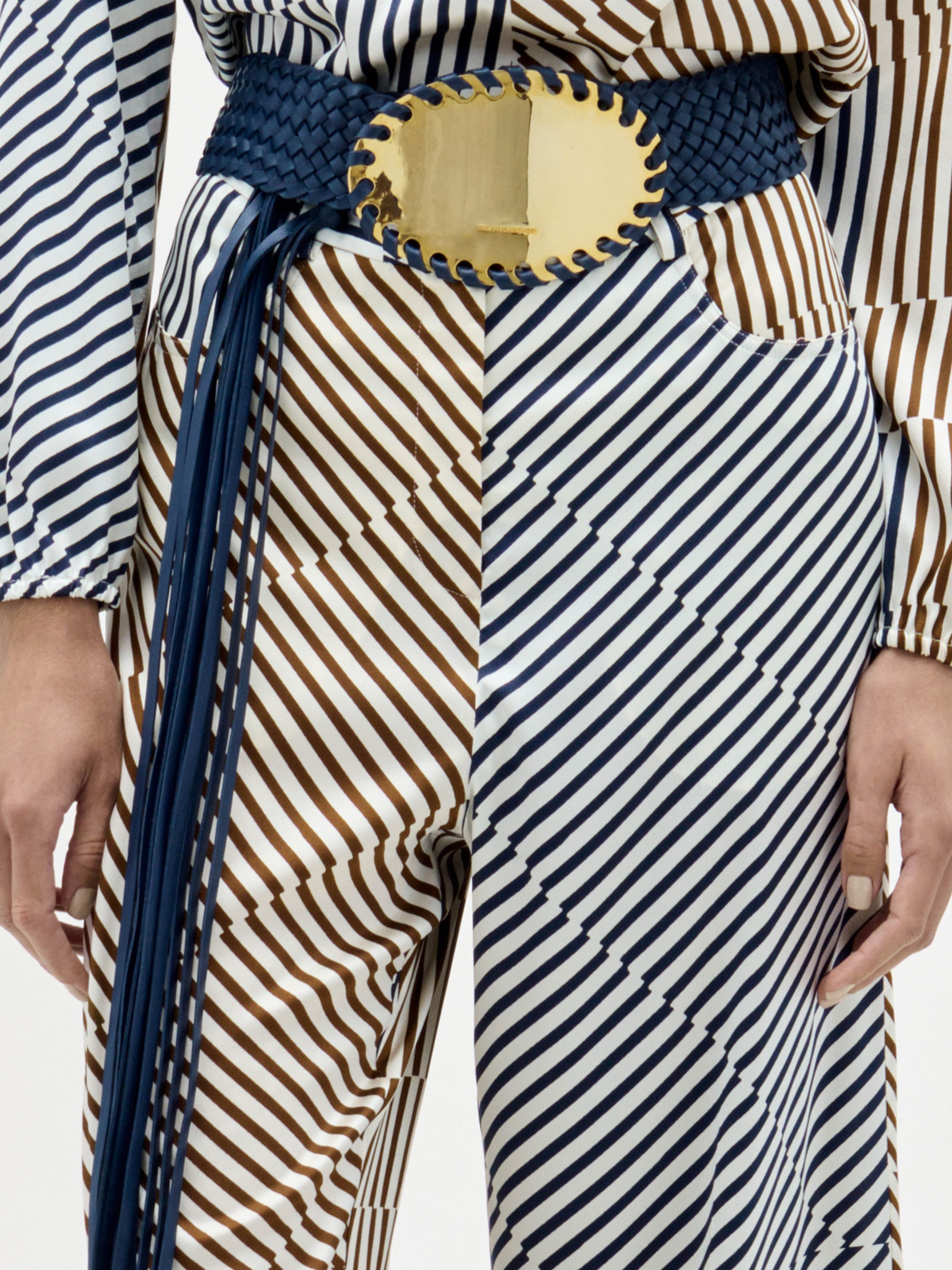 Lleva el pantalón Emine, un conjunto a juego de rayas blancas, marrones y azul marino, combinado con un cinturón ancho azul oscuro con una gran hebilla oval dorada. Solo se ven el torso y la parte superior de las piernas.