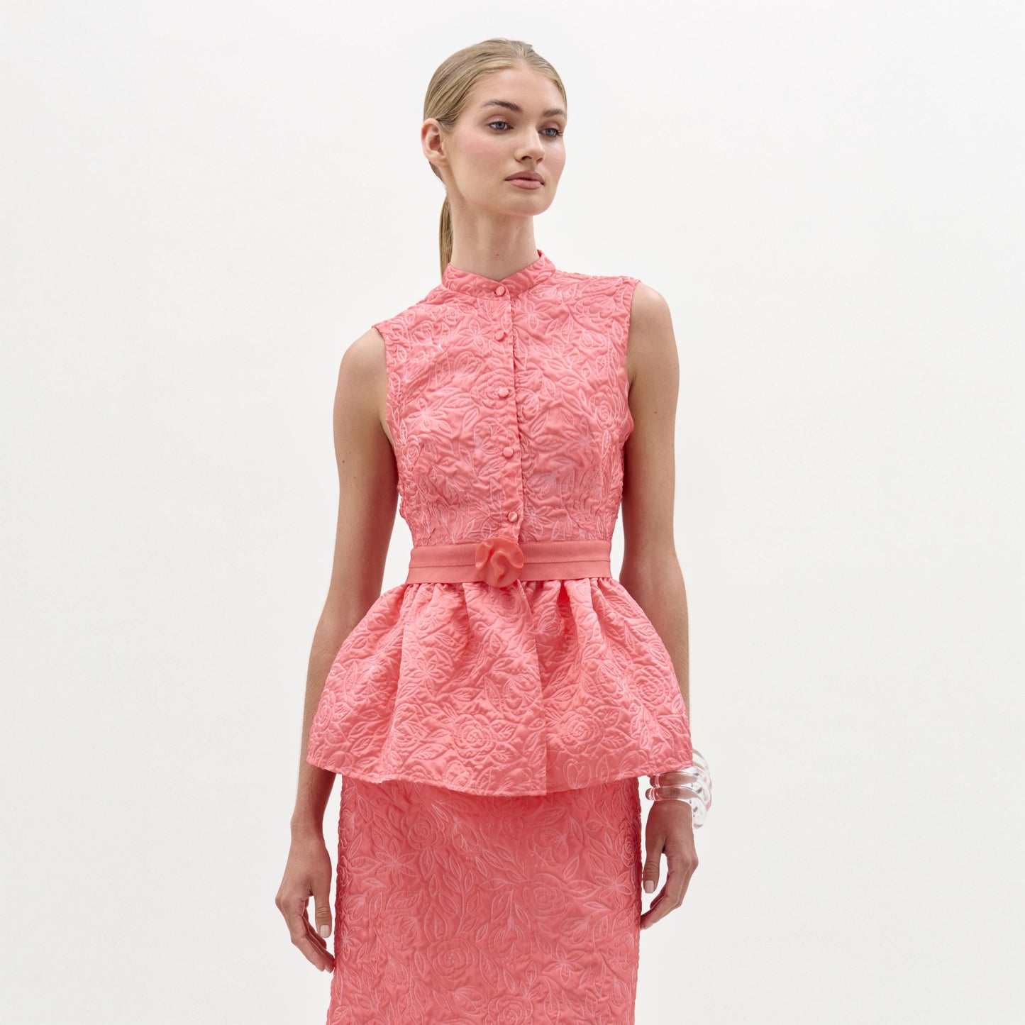 Una mujer de piel clara y pelo rubio liso echado hacia atrás modela la blusa Colony, un top peplum rosa texturizado sin mangas con falda lápiz a juego, fabricado en Colombia, sobre un fondo blanco liso.