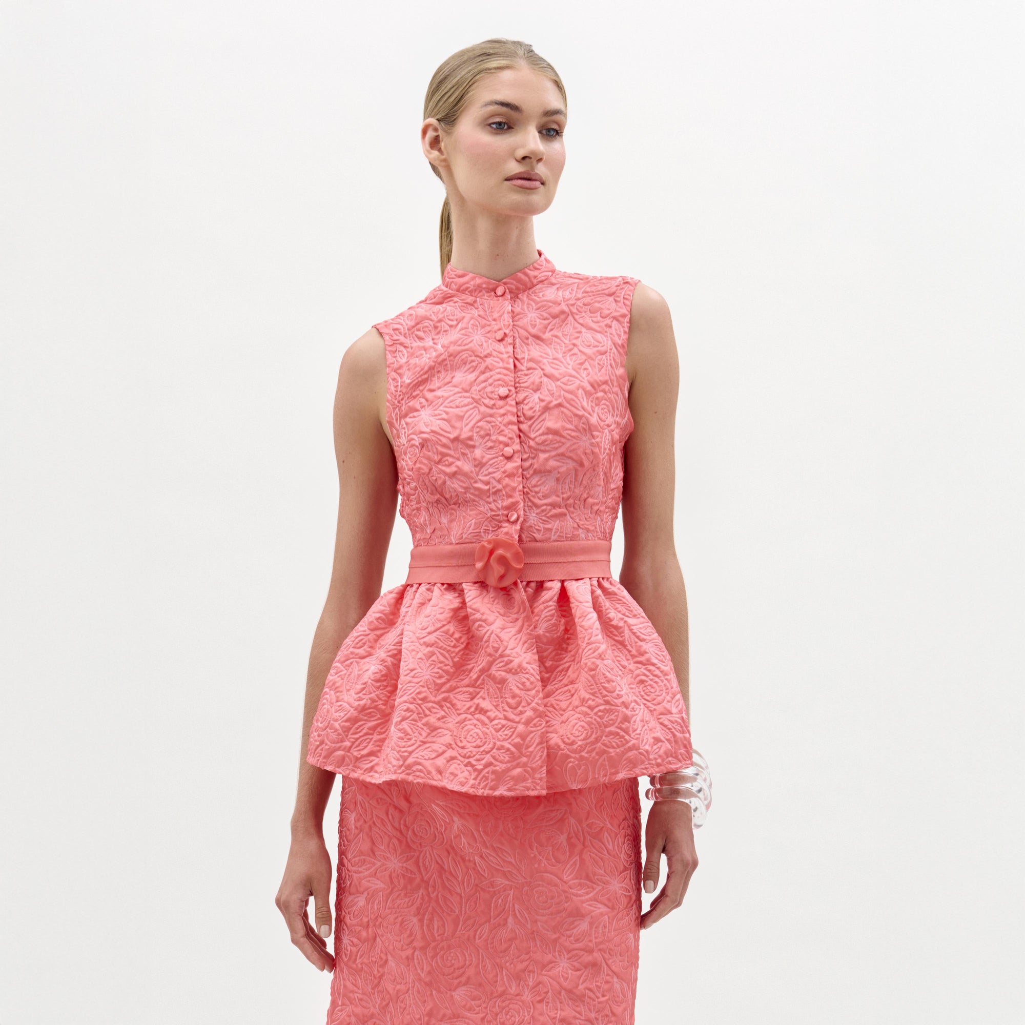 Una mujer de piel clara y pelo rubio liso echado hacia atrás modela la blusa Colony, un top peplum rosa texturizado sin mangas con falda lápiz a juego, fabricado en Colombia, sobre un fondo blanco liso.