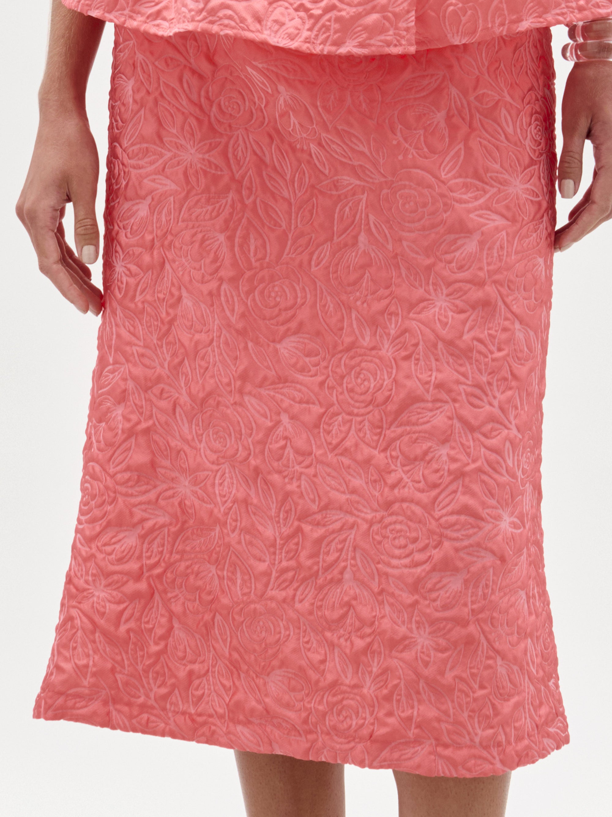 Una persona lleva la falda Atira en rosa coral, una falda midi hasta la rodilla con un estampado floral en relieve confeccionado en tejido italiano. Sus manos están a los lados sobre un fondo blanco liso.