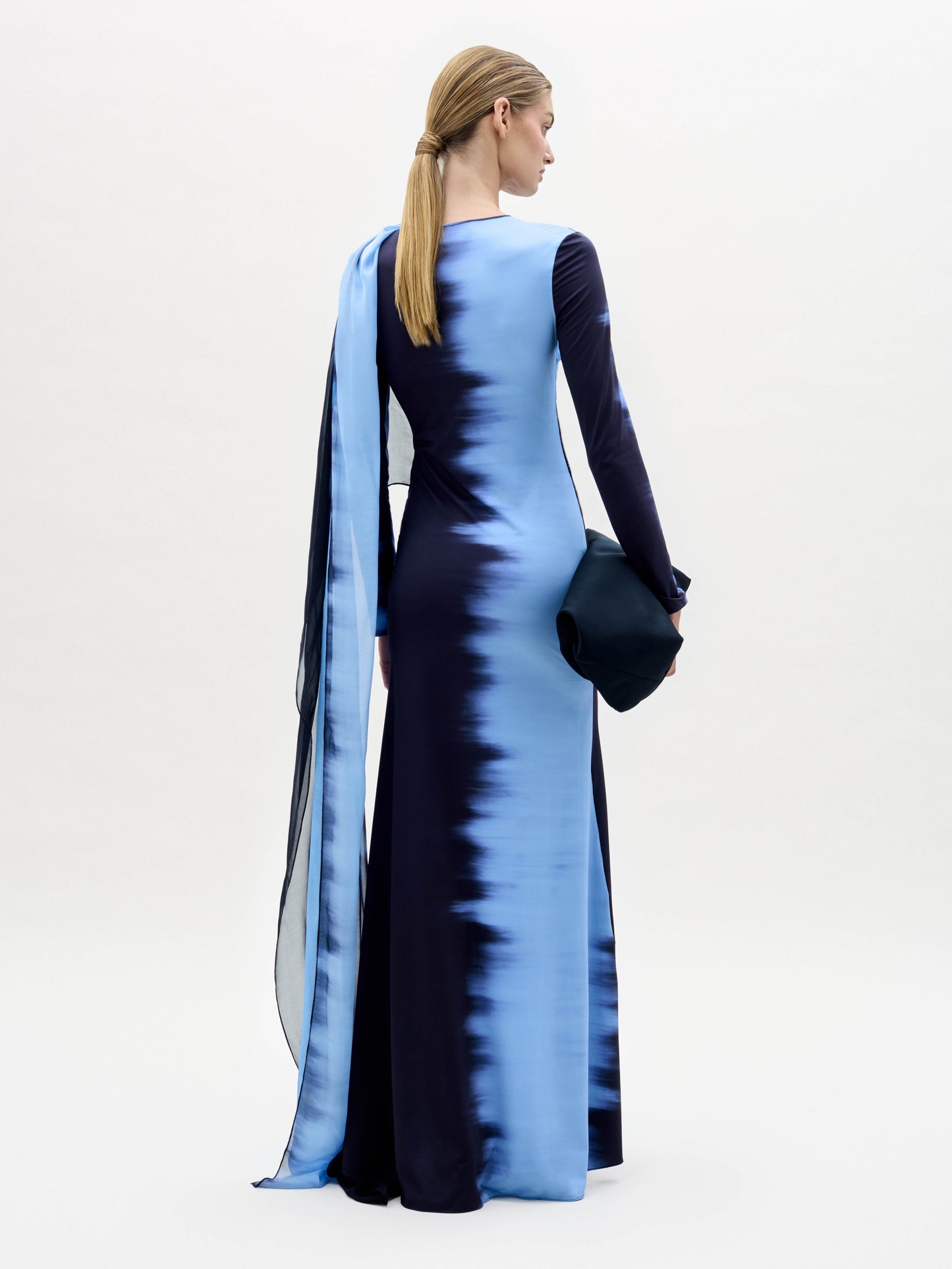 Una persona, de espaldas a nosotros, lleva el Vestido de la Luna, un vestido de manga larga hasta el suelo de color azul oscuro con rayas verticales en degradado azul claro. Lleva un bolso de mano oscuro y el pelo rubio y liso recogido en una coleta baja.