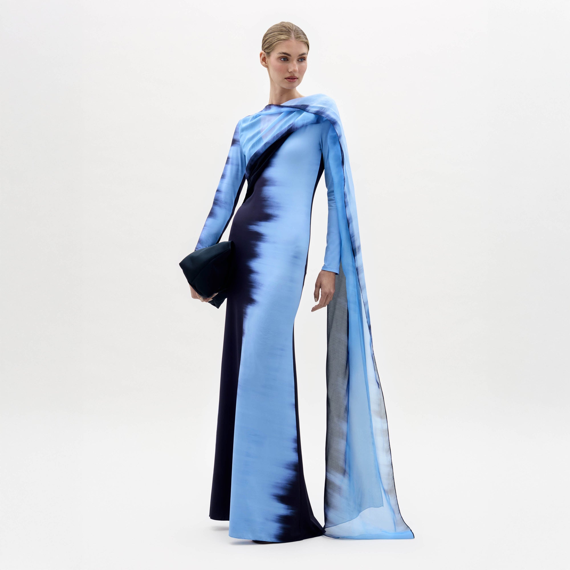 Sobre un fondo liso, una mujer luce el Moon Dress, un vestido largo de manga larga con estampado degradado en azul y negro y detalles en forma de capa, mientras sujeta un bolso de mano negro.