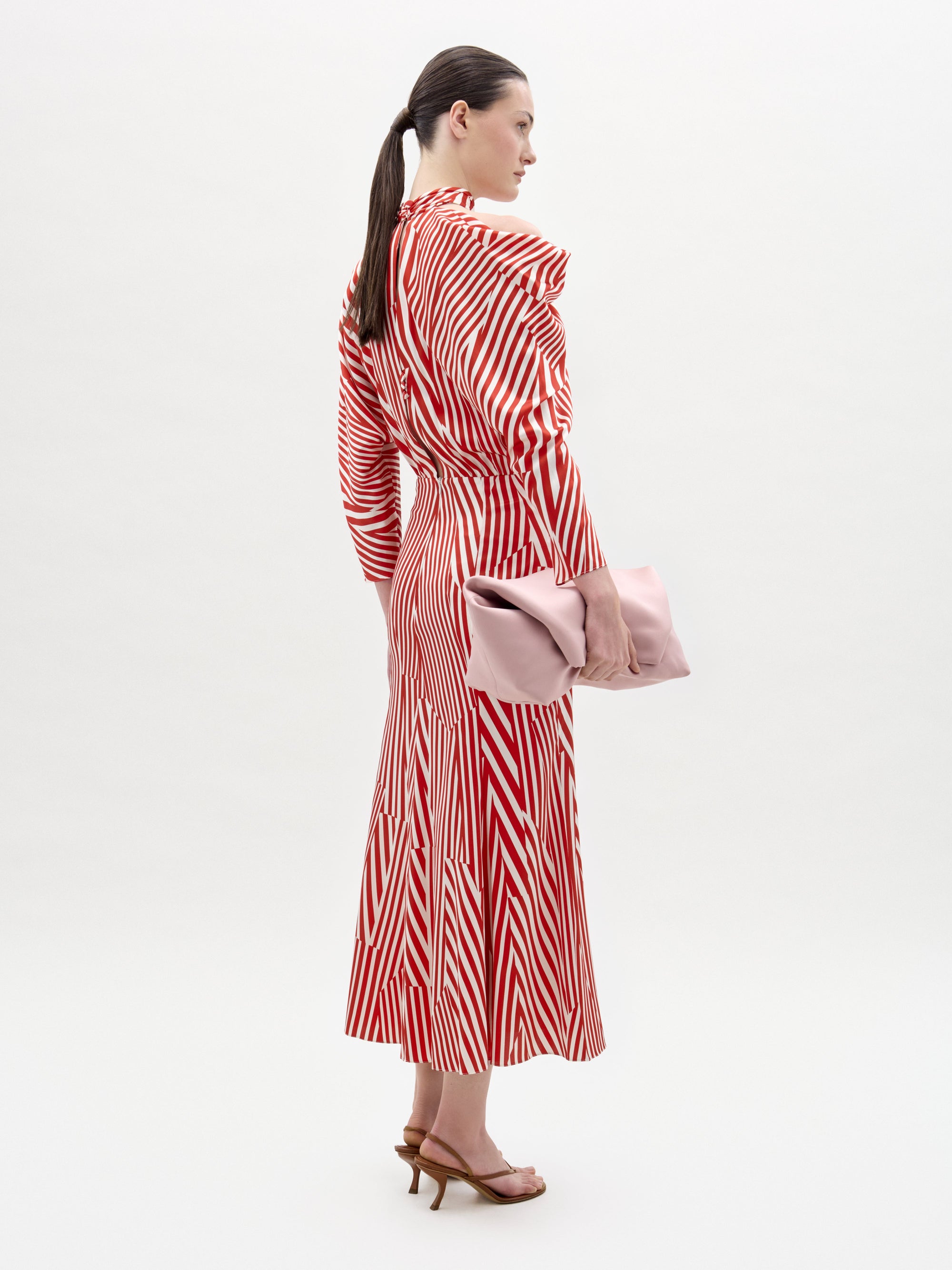 Una mujer, de perfil sobre un fondo liso, lleva el vestido Lisa -un largo midi a rayas rojas y blancas, con cuello alto y mangas largas, confeccionado en tejido italiano- combinado con sandalias de tacón marrones y un gran bolso de mano rosa pálido.