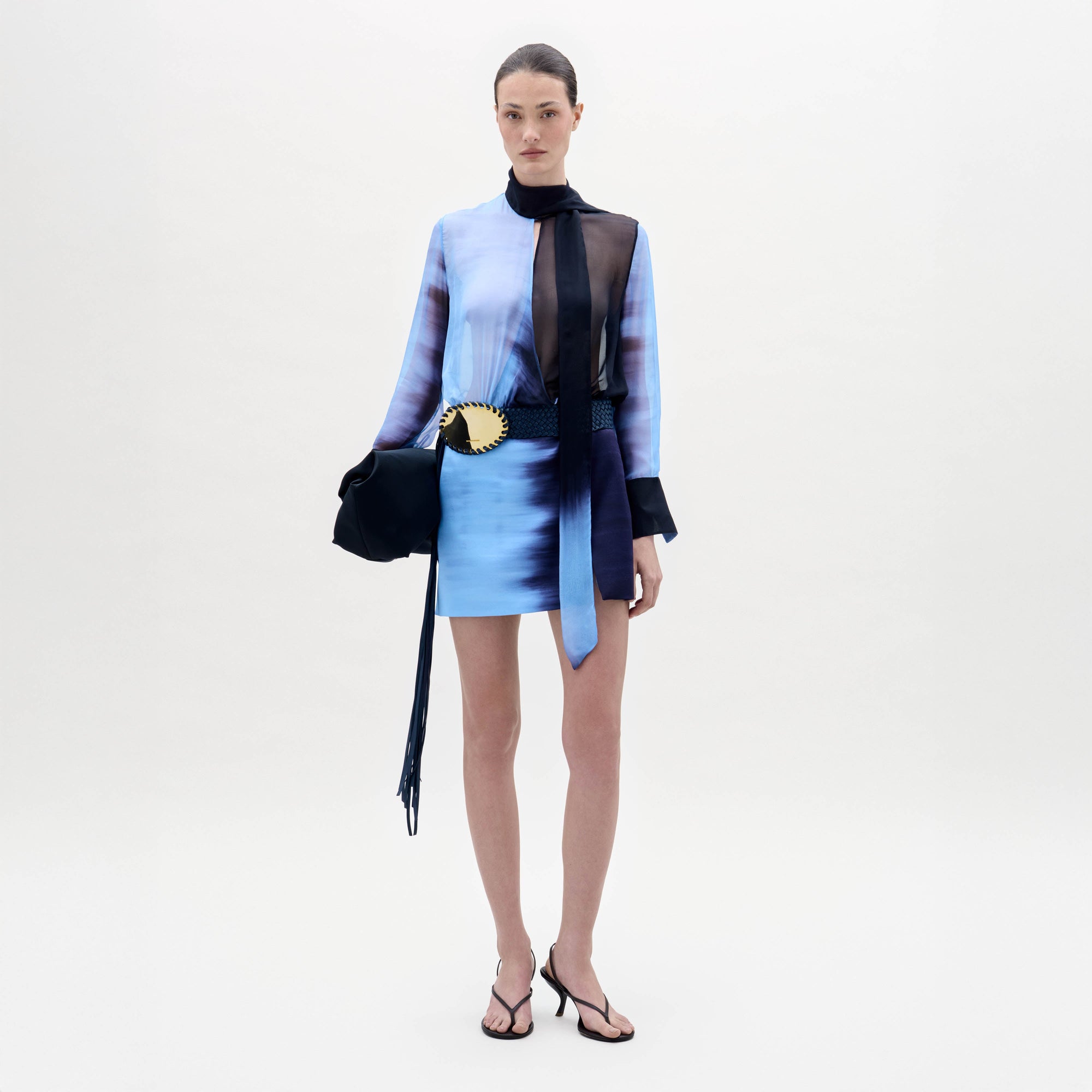 Una mujer posa sobre un fondo blanco con el vestido Imelda, un mini vestido azul degradado con mangas largas transparentes, combinado con un pañuelo negro transparente, un cinturón negro con hebilla de concha dorada, un bolso negro y tacones negros.