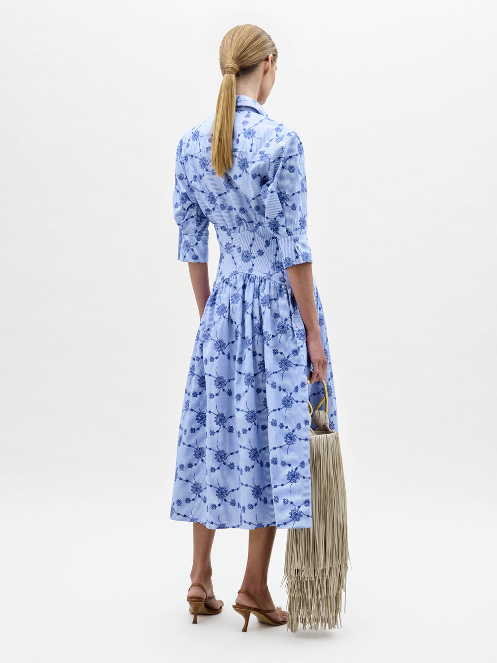 Una mujer con el pelo rubio recogido en una coleta baja, vista de espaldas, lleva el Indy Dress -un vestido azul claro hasta la rodilla con un estampado floral azul oscuro- combinado con un bolso de flecos beige y sandalias de tacón marrones sobre un fondo blanco liso.