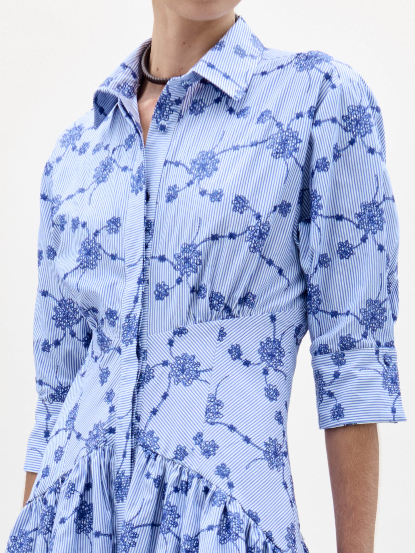 Una persona lleva el Vestido Indy, de rayas verticales azules y blancas, estampado floral azul, mangas tres cuartos y botones delanteros. Su cabeza está parcialmente fuera del encuadre.