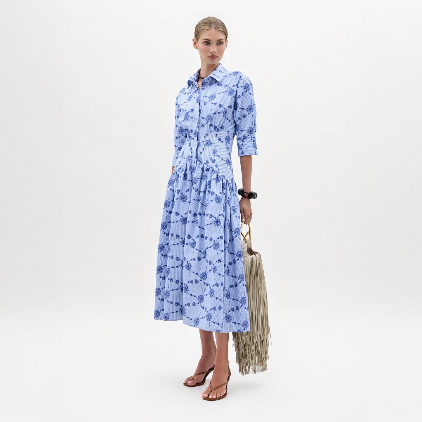 Sobre fondo blanco, una mujer luce el vestido Indy, un vestido midi azul claro estampado con botones y manga tres cuartos. Lo combina con un bolso de mano con flecos y sandalias de tacón marrones.