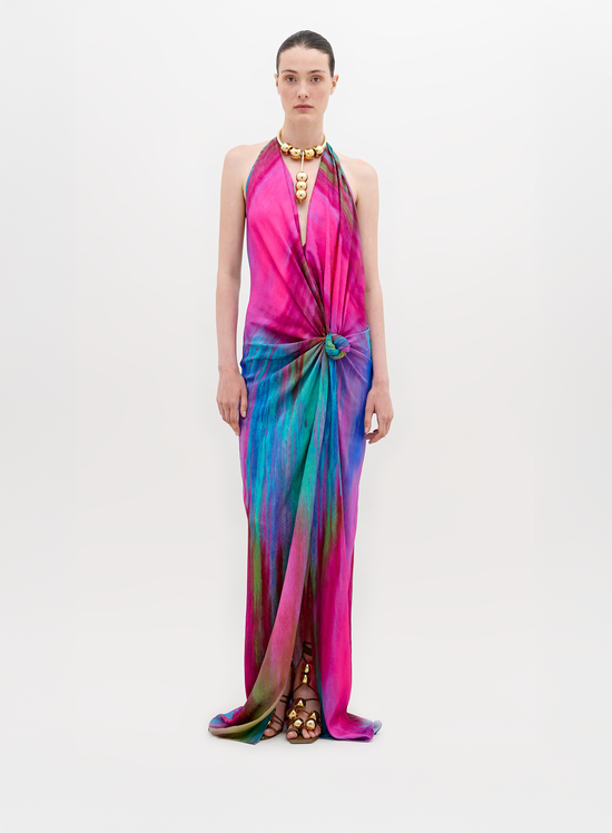 Una persona modela el Vestido Jazmin, un vestido largo fluido en tonos vibrantes de rosa, violeta y azul con efecto tie-dye, con silueta drapeada sobre un fondo blanco liso. Joyas llamativas de oro y sandalias de tiras completan el look.