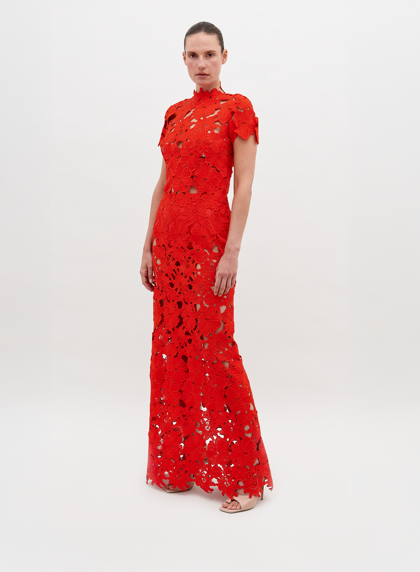 Una persona lleva el Vestido Taiana, un vestido largo de encaje rojo brillante con estampados florales. La silueta ajustada incluye mangas cortas y cuello alto. Esta exquisita creación de Vestido Atelier se destaca sobre un fondo blanco liso.