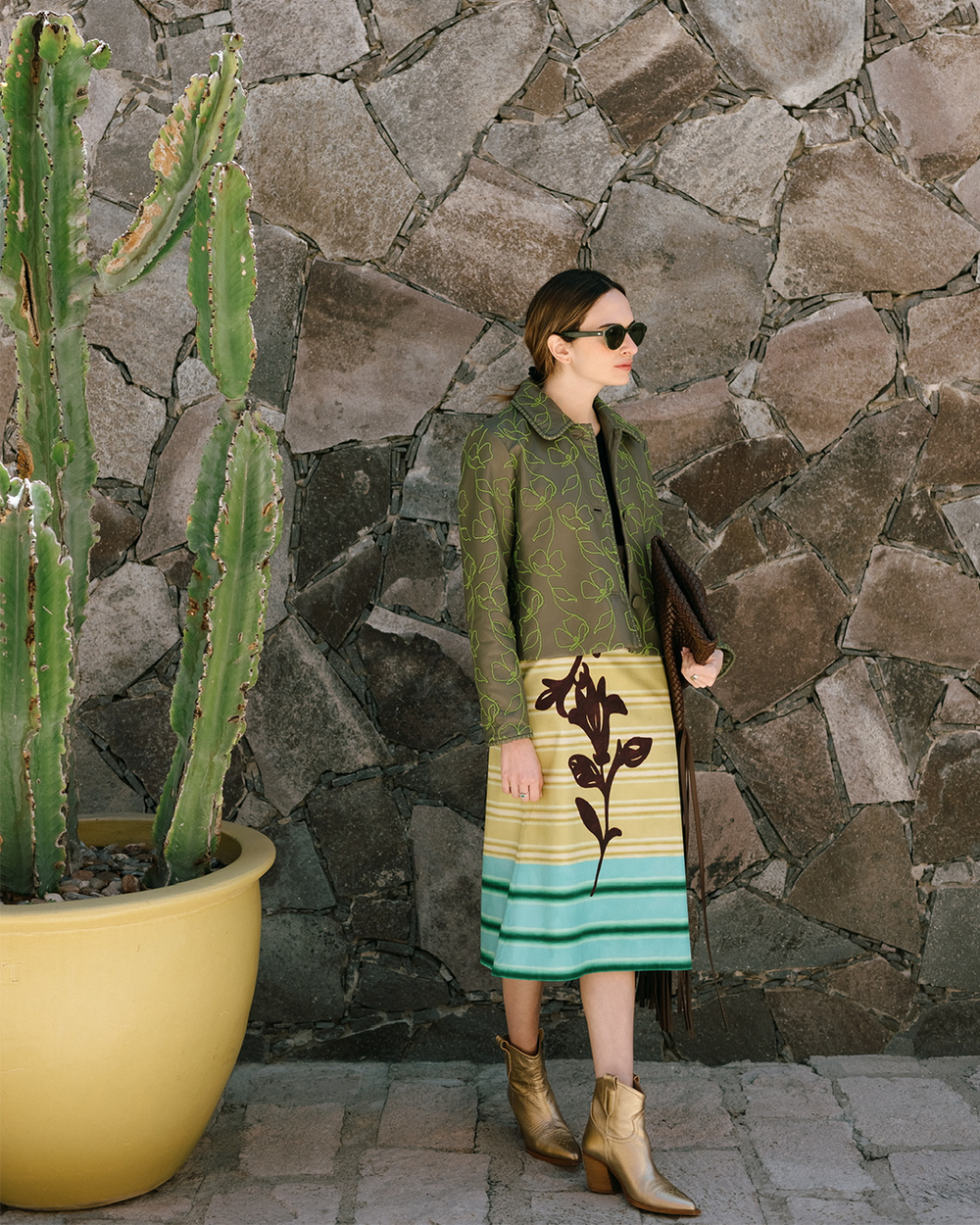 Una persona se encuentra al aire libre junto a un gran cactus en maceta contra un muro de piedra. Lleva gafas de sol, un abrigo verde con un diseño floral, una falda con rayas horizontales verdes y amarillas y botines marrones, mientras sostiene un bolso estampado.