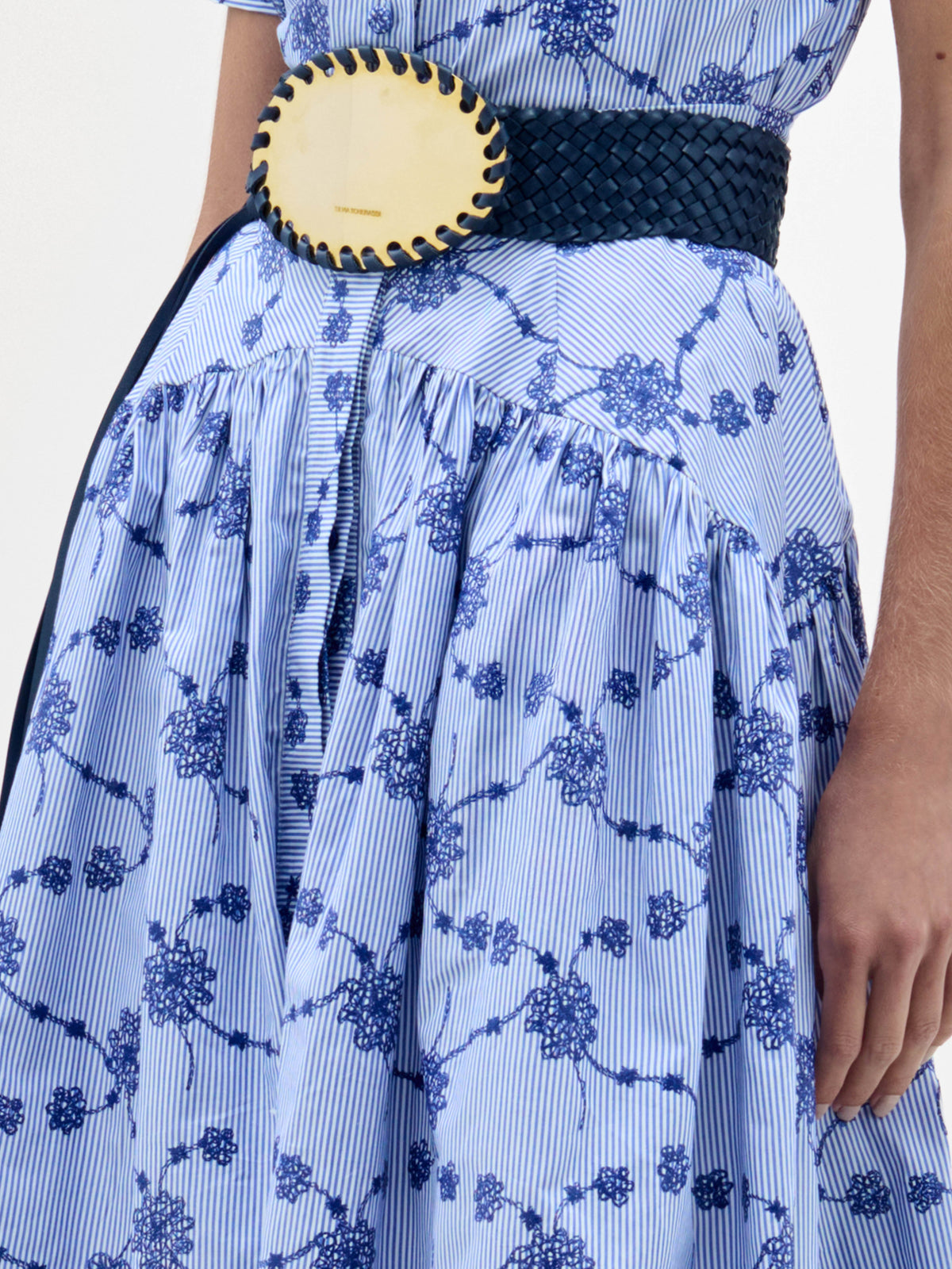 Lleva la falda Narel, de rayas azules y blancas con motivos florales, con un cinturón ancho de tejido negro con una gran hebilla oval cosida. Sólo se ven el torso y el brazo derecho.