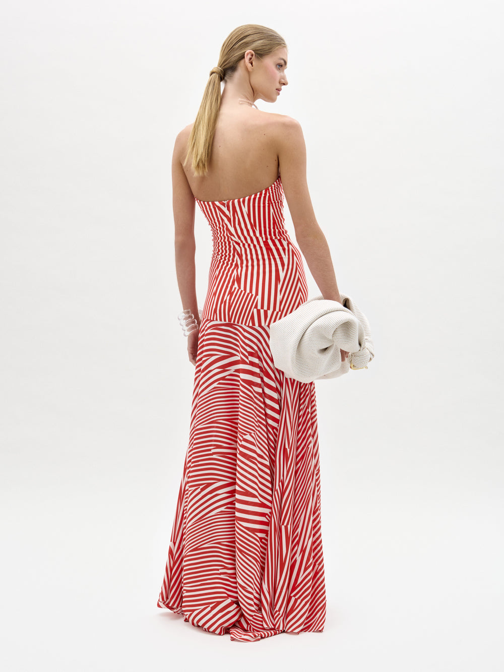 Una mujer mira hacia otro lado con el vestido Muriel, un maxi sin tirantes de tela italiana con rayas rojas y blancas. Lleva un bolso de mano grande y ligero y el pelo rubio y liso recogido. Sobre un fondo blanco liso. Fabricado en Colombia.