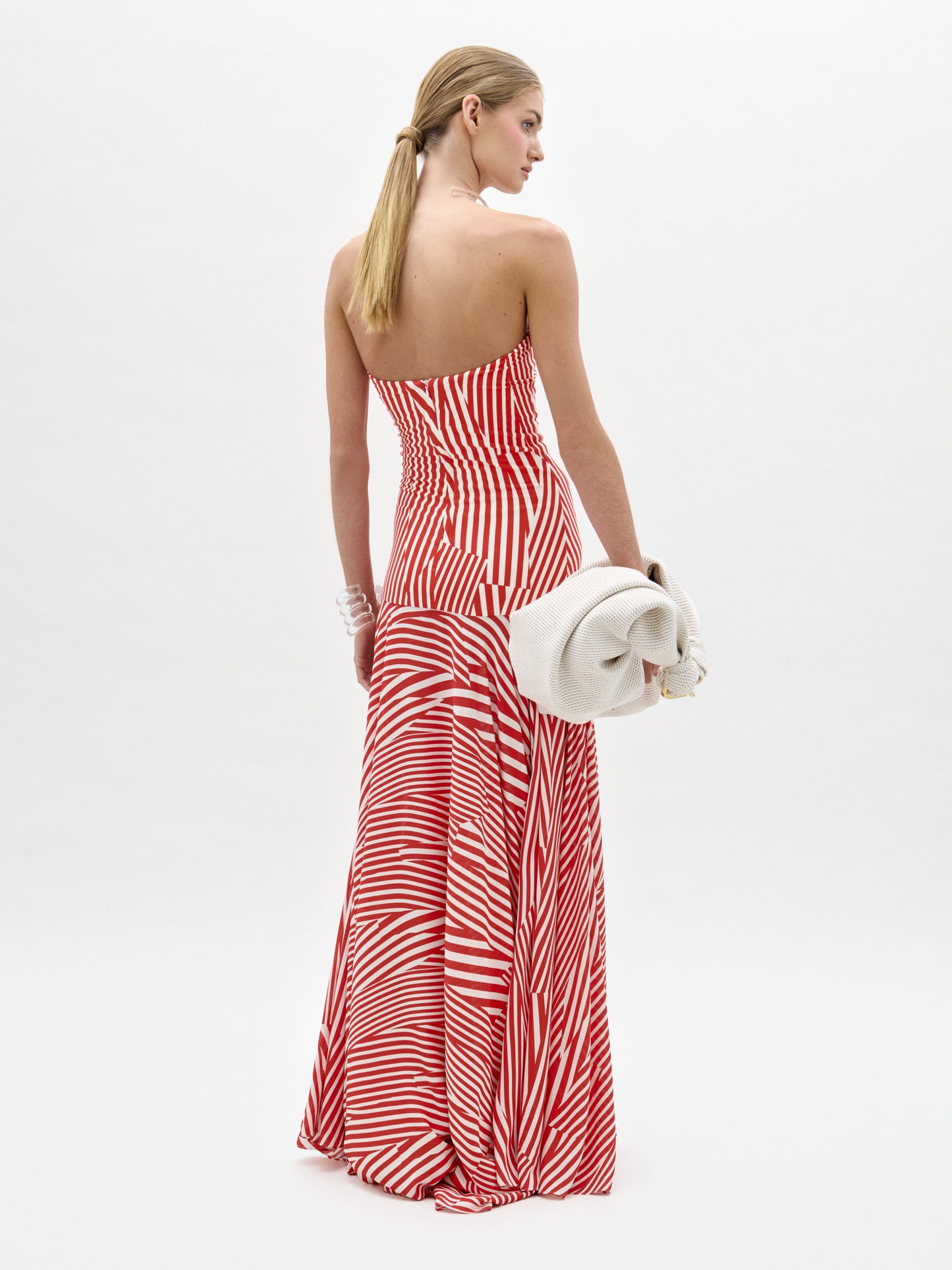 Una mujer mira hacia otro lado con el vestido Muriel, un maxi sin tirantes de tela italiana con rayas rojas y blancas. Lleva un bolso de mano grande y ligero y el pelo rubio y liso recogido. Sobre un fondo blanco liso. Fabricado en Colombia.