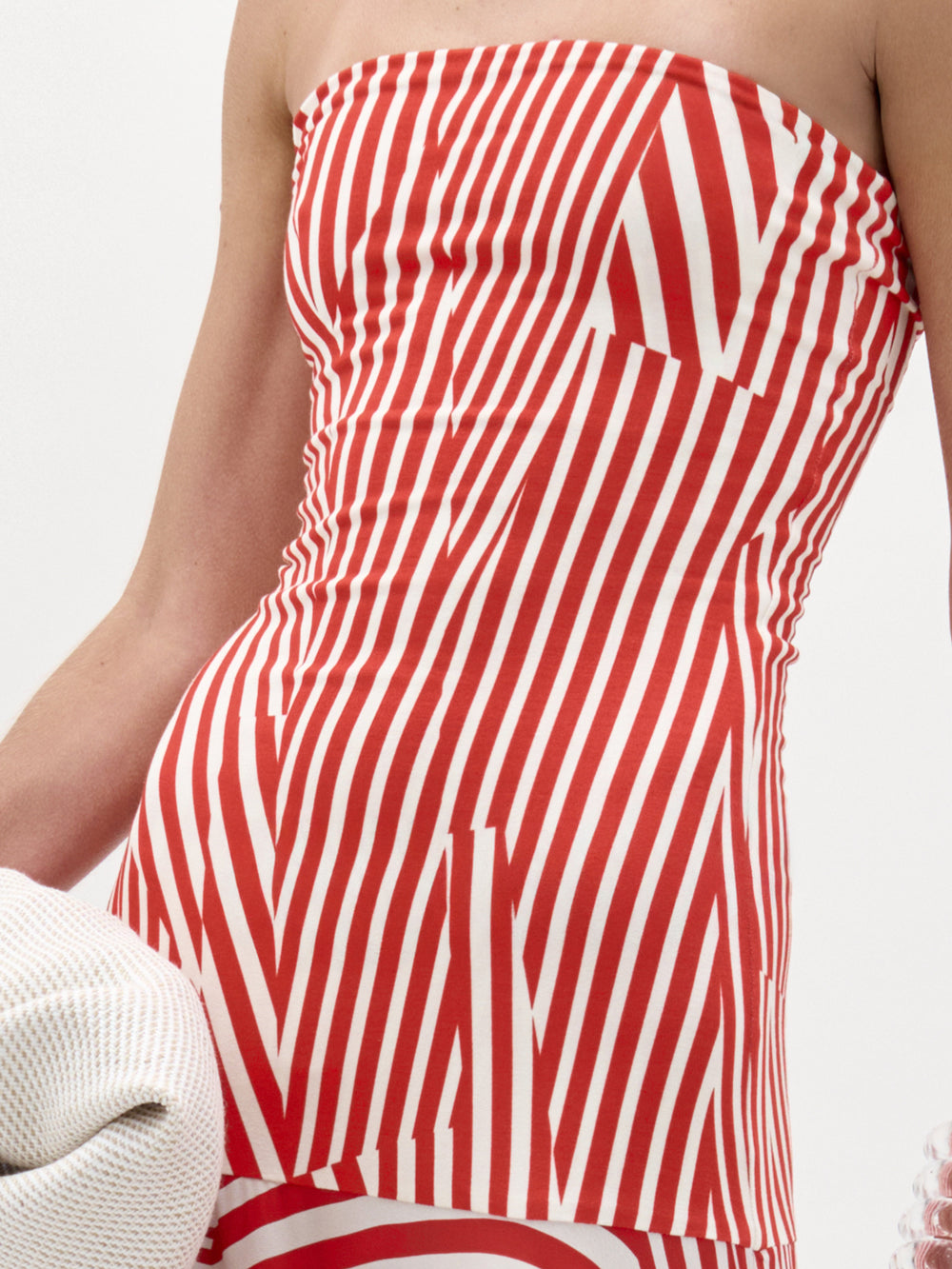 Una persona modela el Vestido Muriel -un diseño maxi sin tirantes a rayas rojas y blancas con líneas abstractas verticales y diagonales- de pie sobre un fondo liso, con parte de su brazo y un objeto blanco texturizado visible.