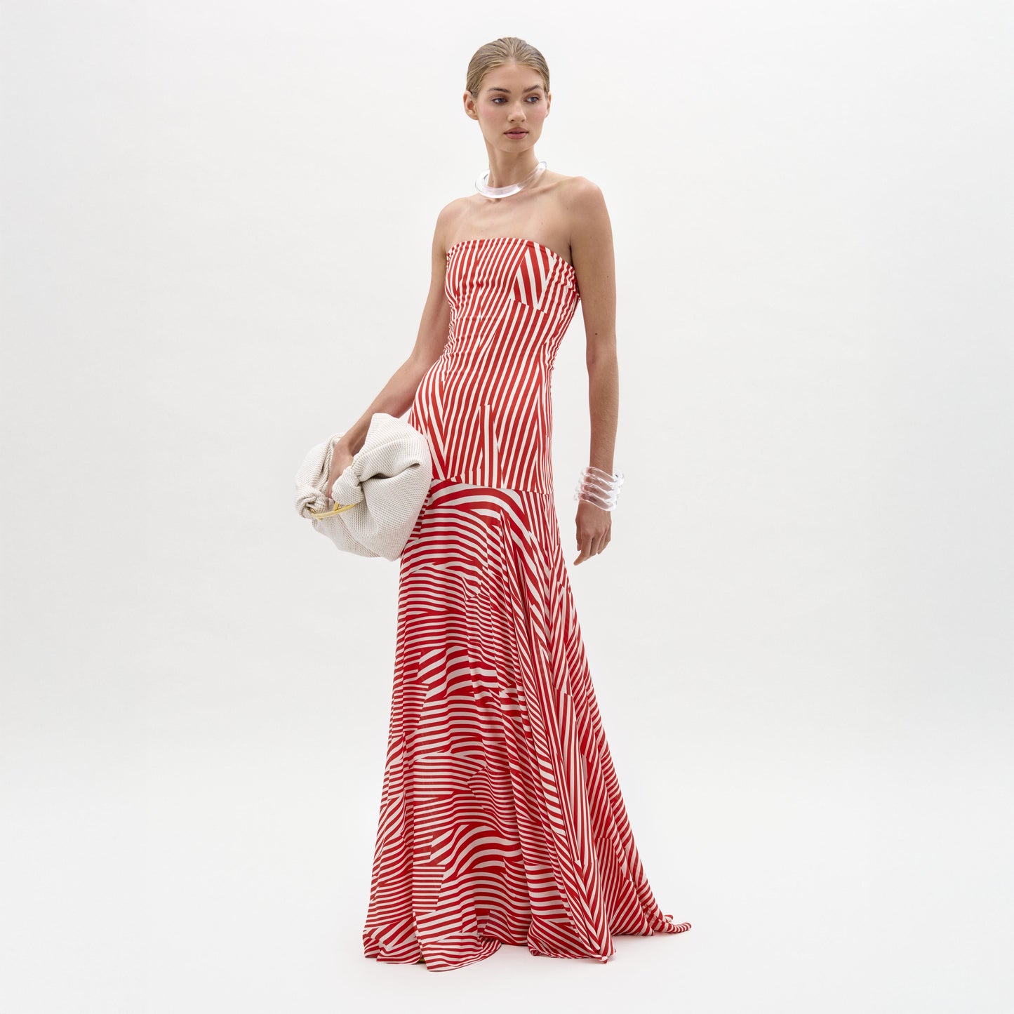 Sobre un fondo blanco liso, una persona luce el vestido Muriel, sin tirantes, a rayas rojas y blancas, hecho en Colombia, con un bolso de mano blanco, un collar de perlas y pulseras.
