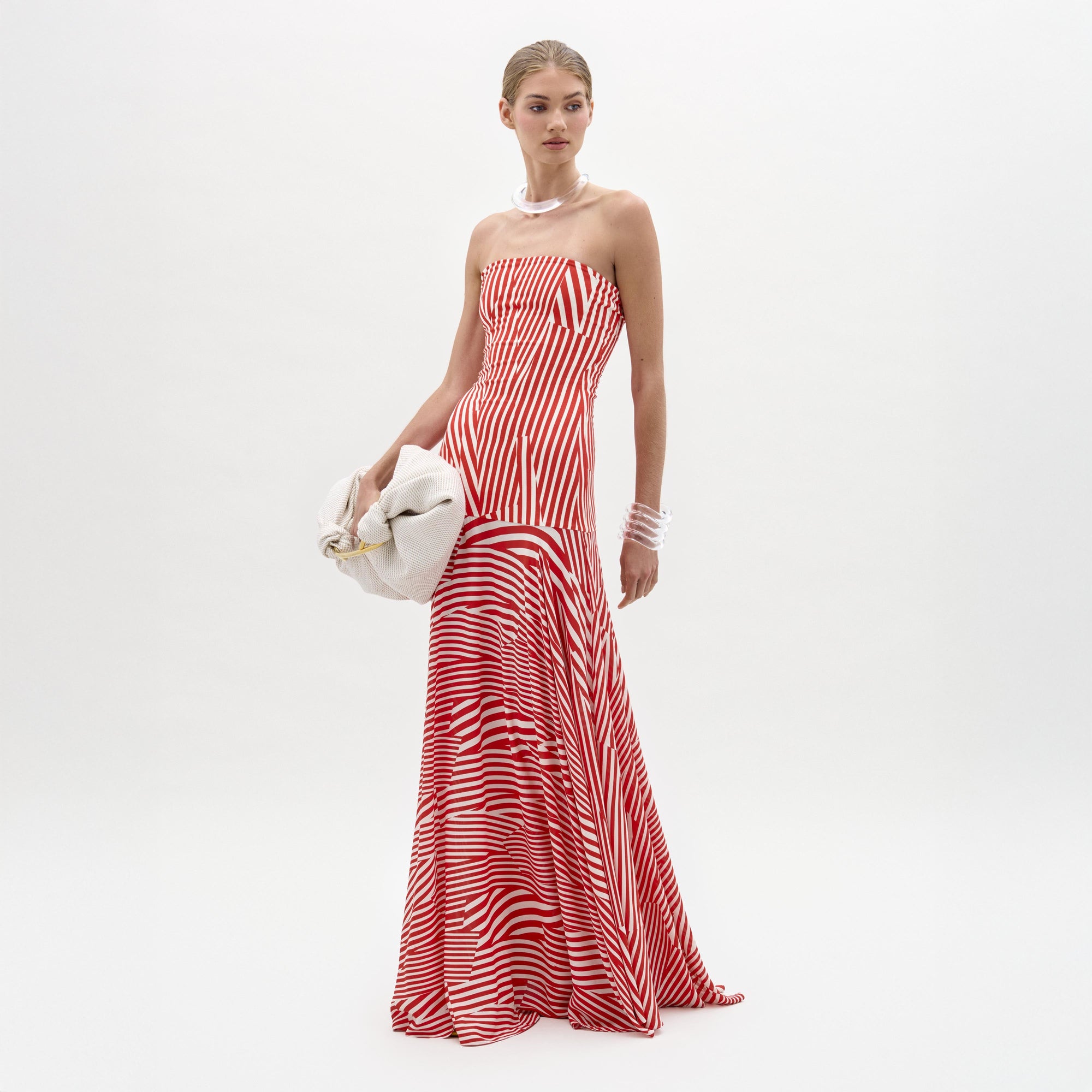Sobre un fondo blanco liso, una persona luce el vestido Muriel, sin tirantes, a rayas rojas y blancas, hecho en Colombia, con un bolso de mano blanco, un collar de perlas y pulseras.
