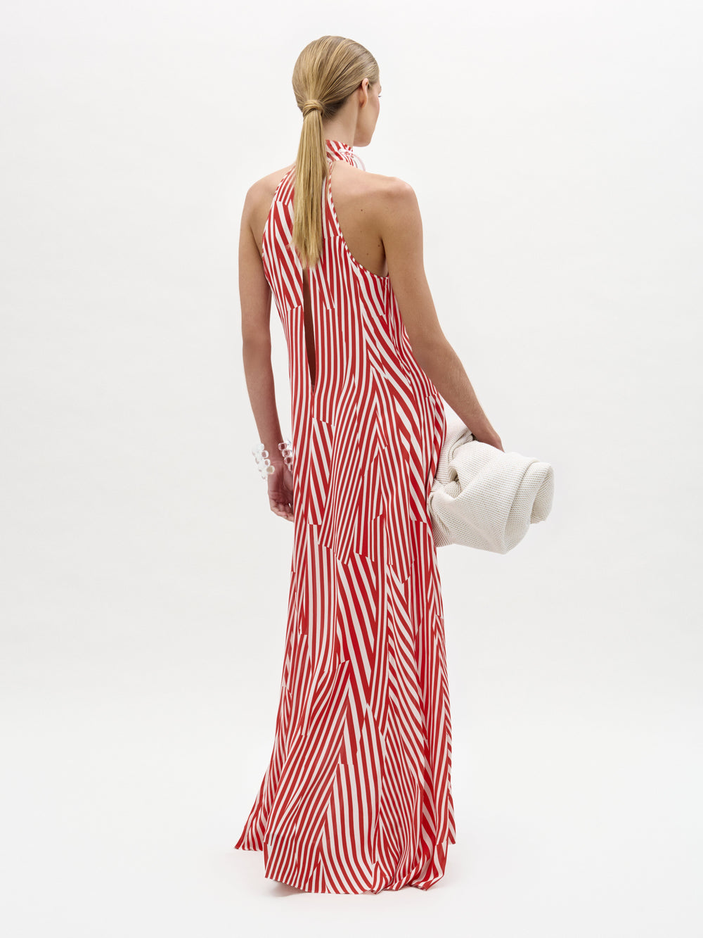 Una persona con el pelo rubio, largo y liso, de pie y de espaldas, lleva el vestido sin mangas de rayas rojas y blancas Koa Dress, fabricado en Colombia, y sostiene una toalla blanca enrollada sobre un fondo liso y claro.