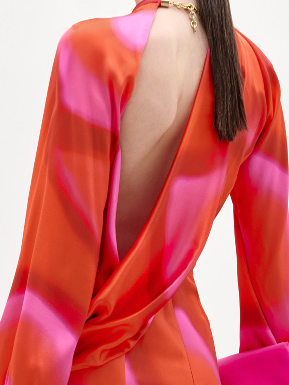 Una persona con el pelo liso y oscuro lleva el vestido Muna -un midi de seda italiana con estampado rojo y rosa y espalda descubierta- de manga larga y un collar de cadena dorada, que cae con elegancia y deja al descubierto parte de la piel.