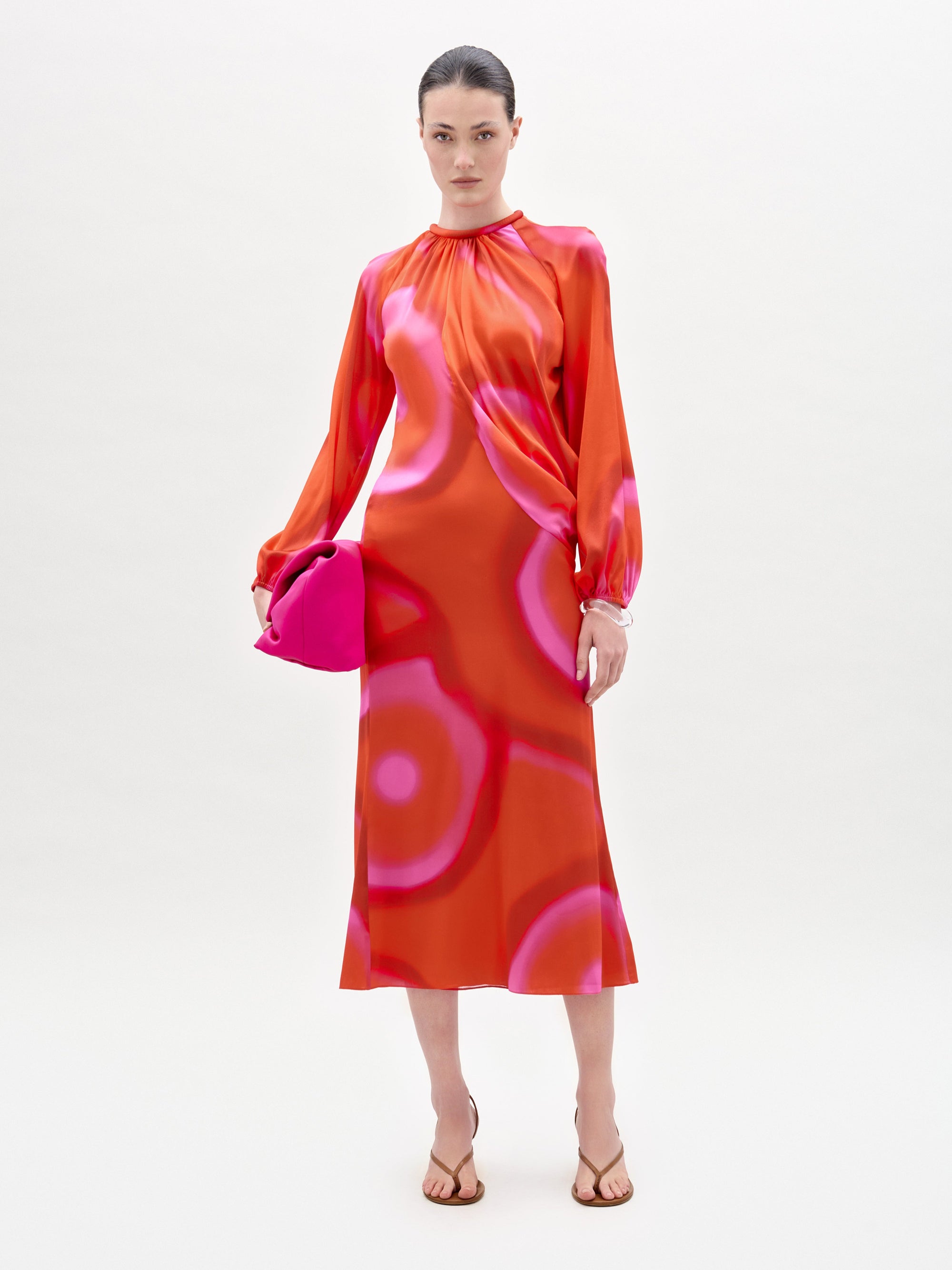 Una mujer, sobre un fondo claro y liso, lleva el vestido Muna, una falda tobillera de seda italiana con motivos circulares rosas, un bolso de mano rosa brillante y sandalias beige.