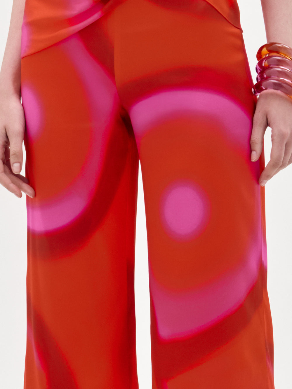 Una persona lleva el pantalón Andie Pant en naranja brillante con grandes círculos rosas y corte de pernera ancha, peinado con gruesos brazaletes naranja translúcido en un brazo, sobre un fondo blanco liso.