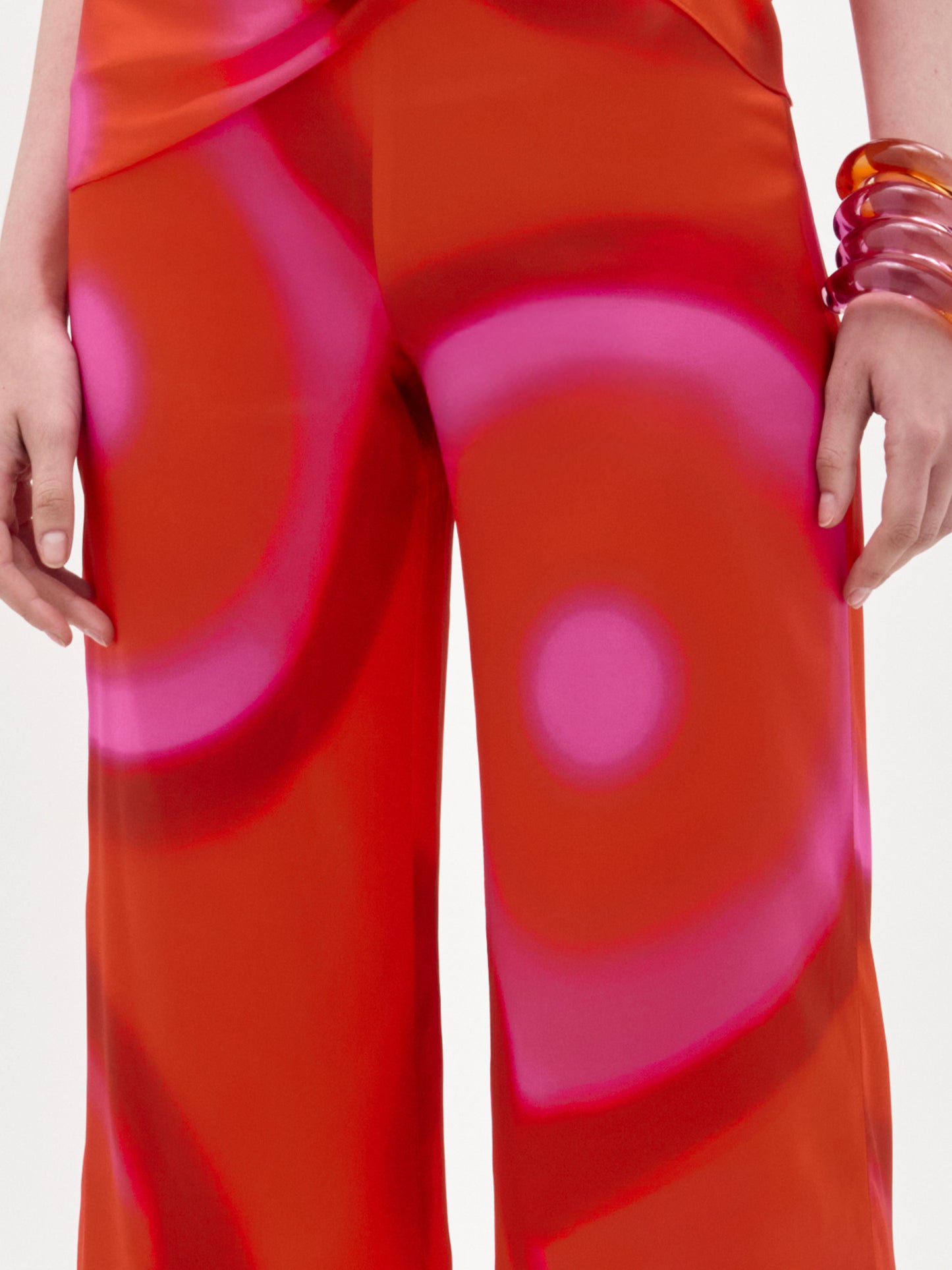 Una persona lleva el pantalón Andie Pant en naranja brillante con grandes círculos rosas y corte de pernera ancha, peinado con gruesos brazaletes naranja translúcido en un brazo, sobre un fondo blanco liso.