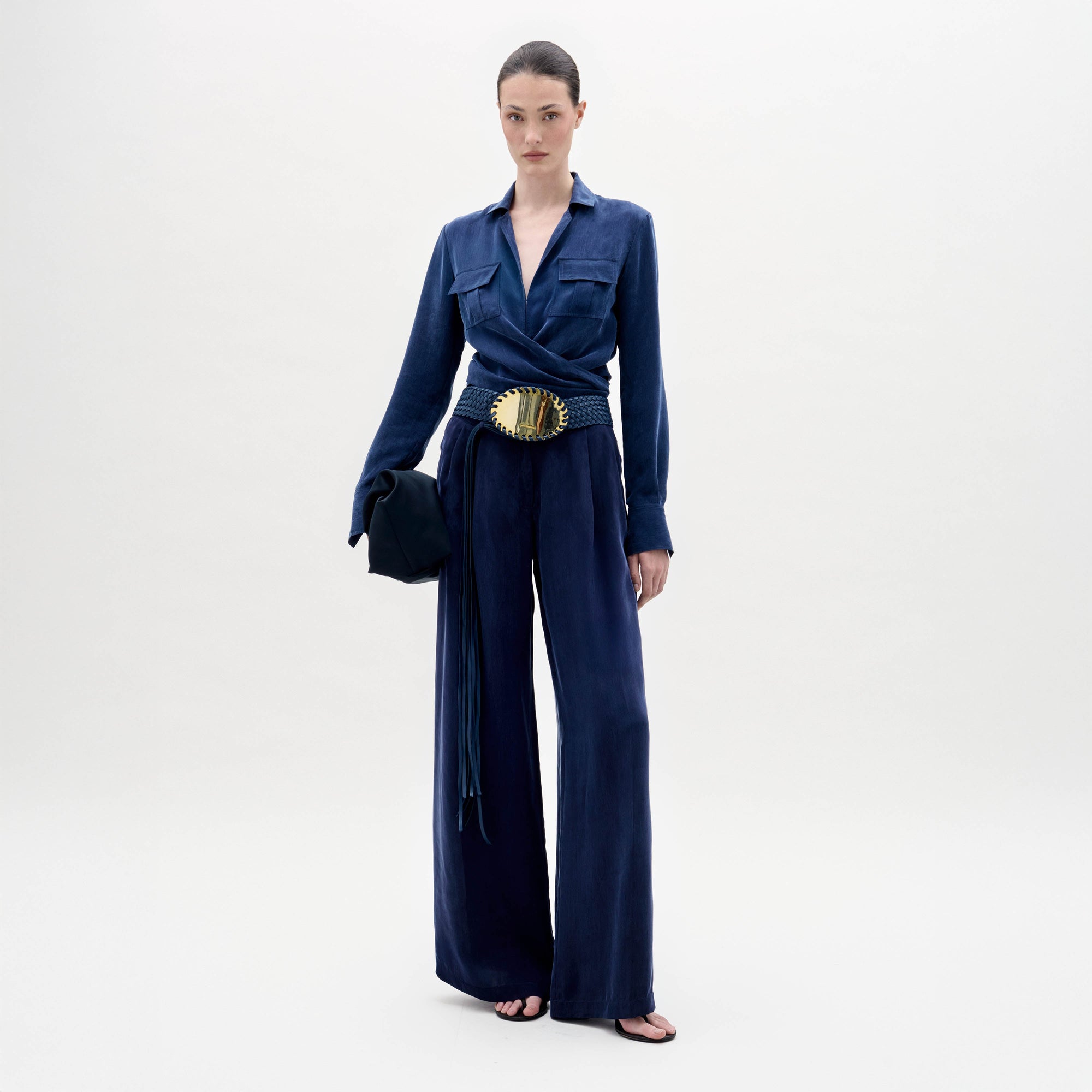 Una persona de pie sobre un fondo claro liso lleva una blusa azul oscuro de manga larga, pantalones de pata ancha Opal Pant, un cinturón negro y dorado, sandalias negras y un bolso negro en la mano.