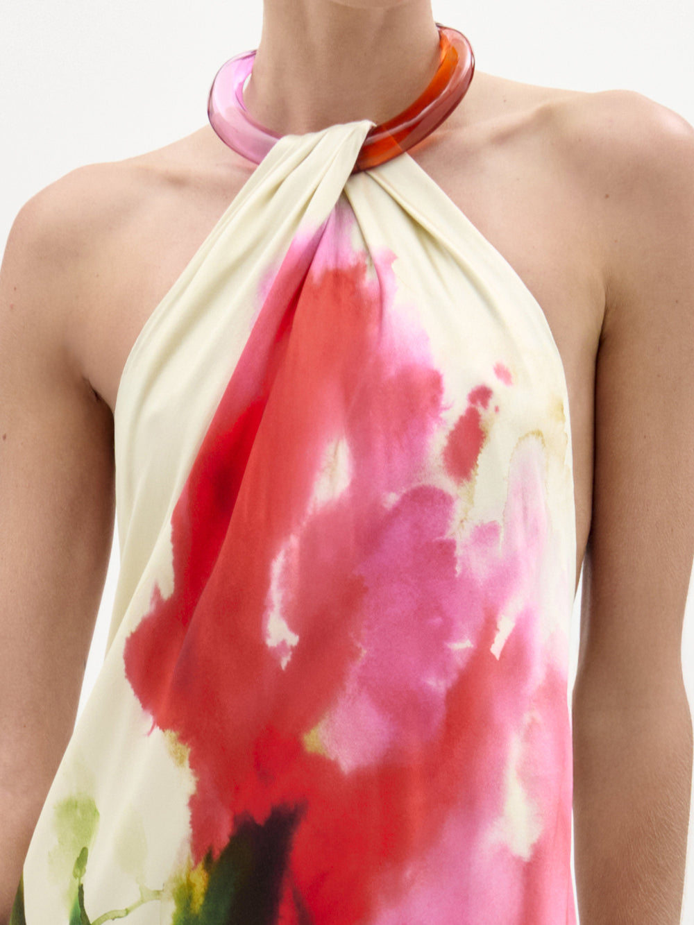 Una persona lleva el Jesse Dress, un maxi halter sin mangas con un estampado de acuarelas rosas y rojas, combinado con un collar circular translúcido de gran tamaño en rosa y naranja. Fabricado en Colombia, destaca sobre un fondo liso y claro.