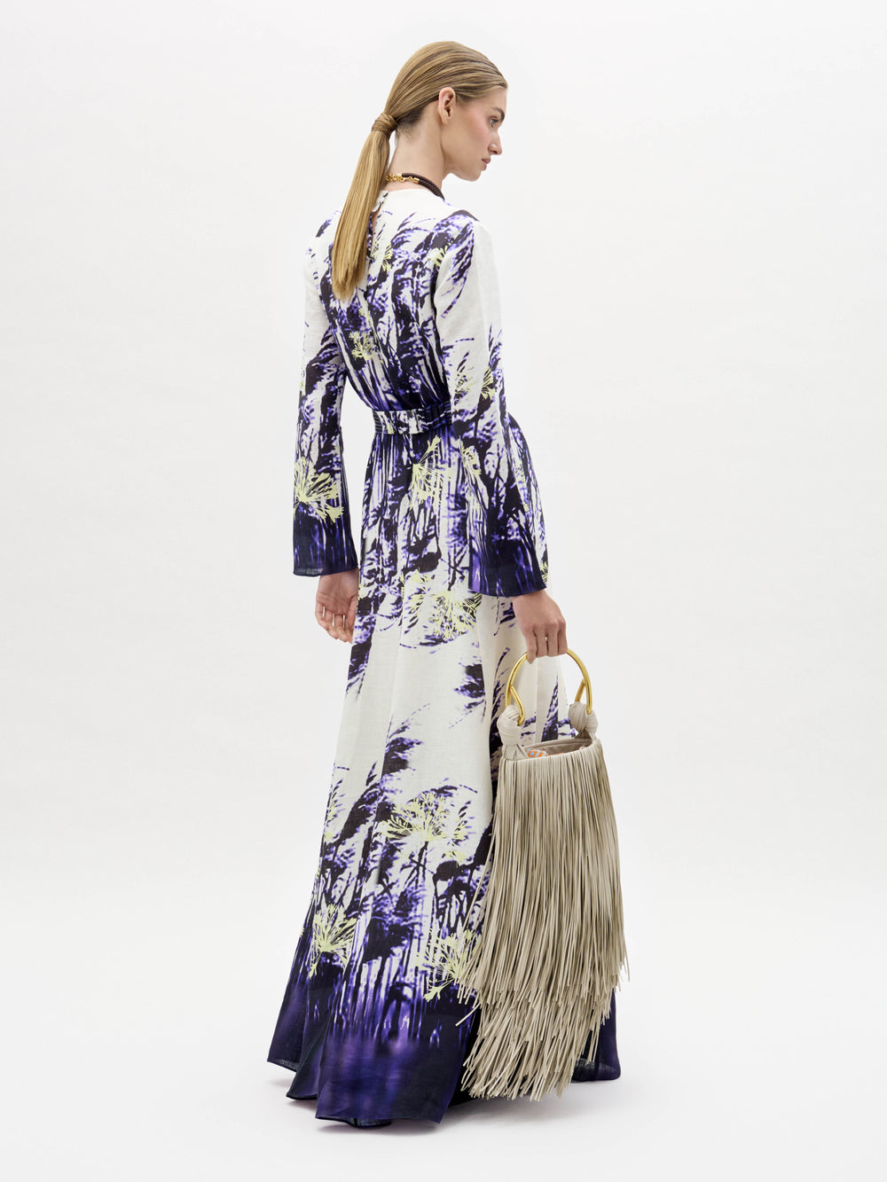 Una mujer, de perfil, lleva el vestido Kady, largo hasta el suelo y de manga larga, con estampado de naturaleza en morado y blanco. Lleva un bolso grande de flecos beige con asas circulares y el pelo liso recogido.