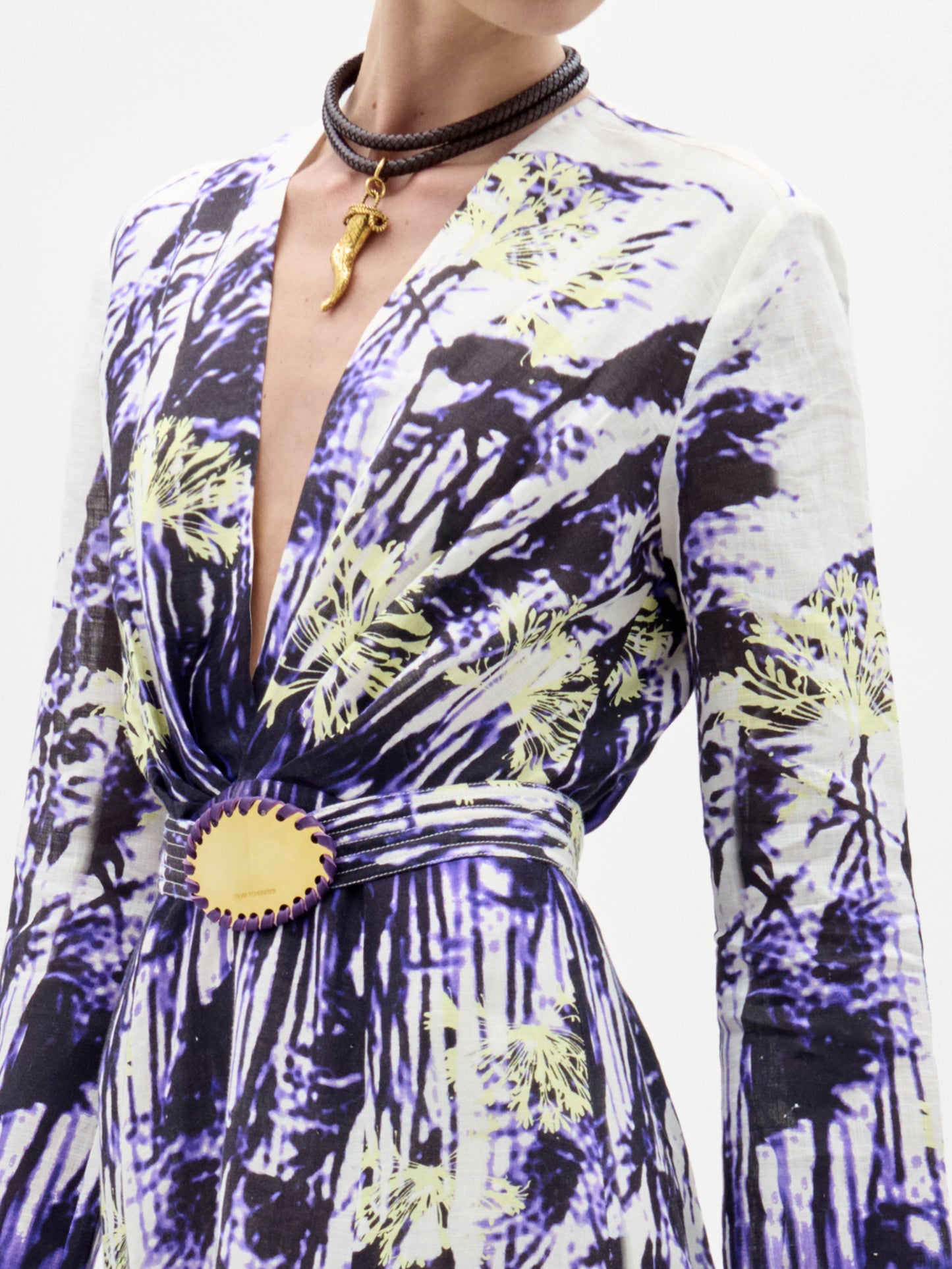 Una persona lleva el vestido Kady, con un profundo escote en V y estampado floral abstracto azul y amarillo, combinado con un cinturón a juego con hebilla ovalada amarilla y un collar gargantilla marrón adornado con un colgante dorado.
