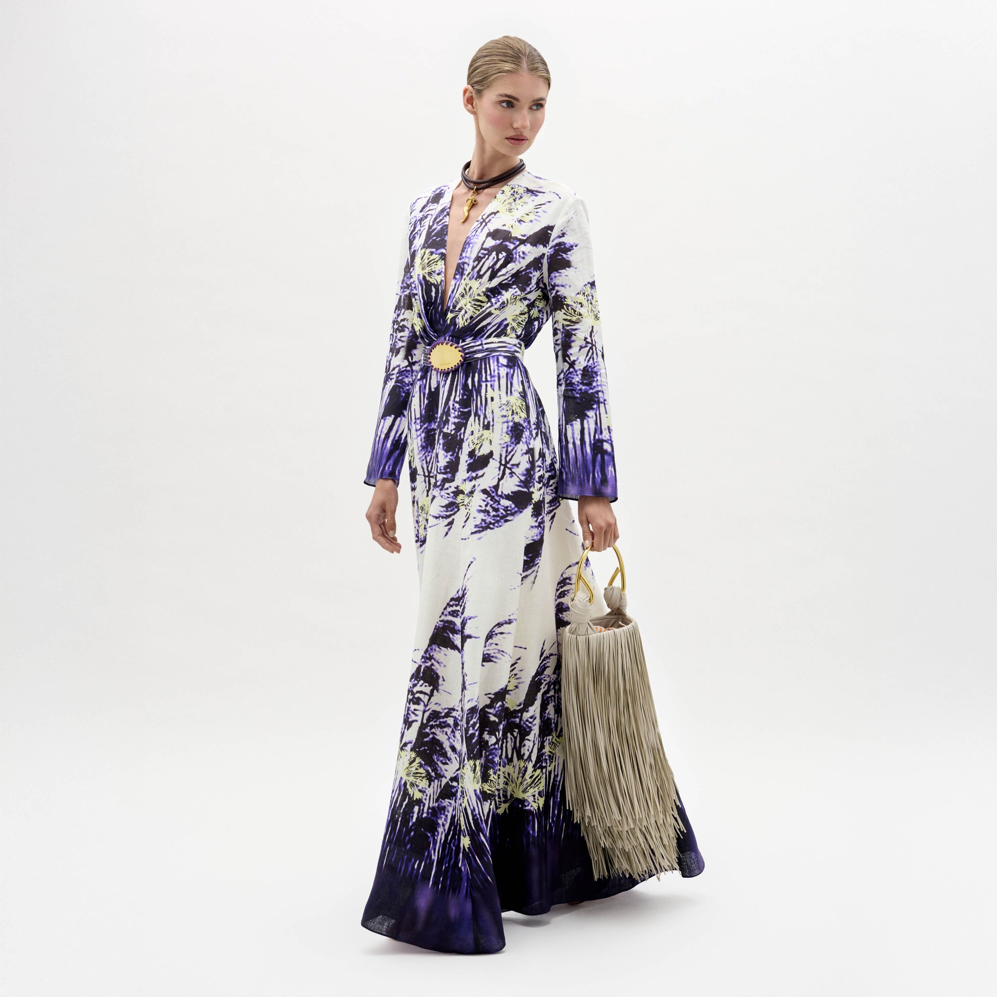 Una mujer sobre fondo blanco luce el vestido Kady, una prenda larga estampada con motivos morados y verdes, combinada con un cinturón, una gargantilla y un bolso grande con flecos.