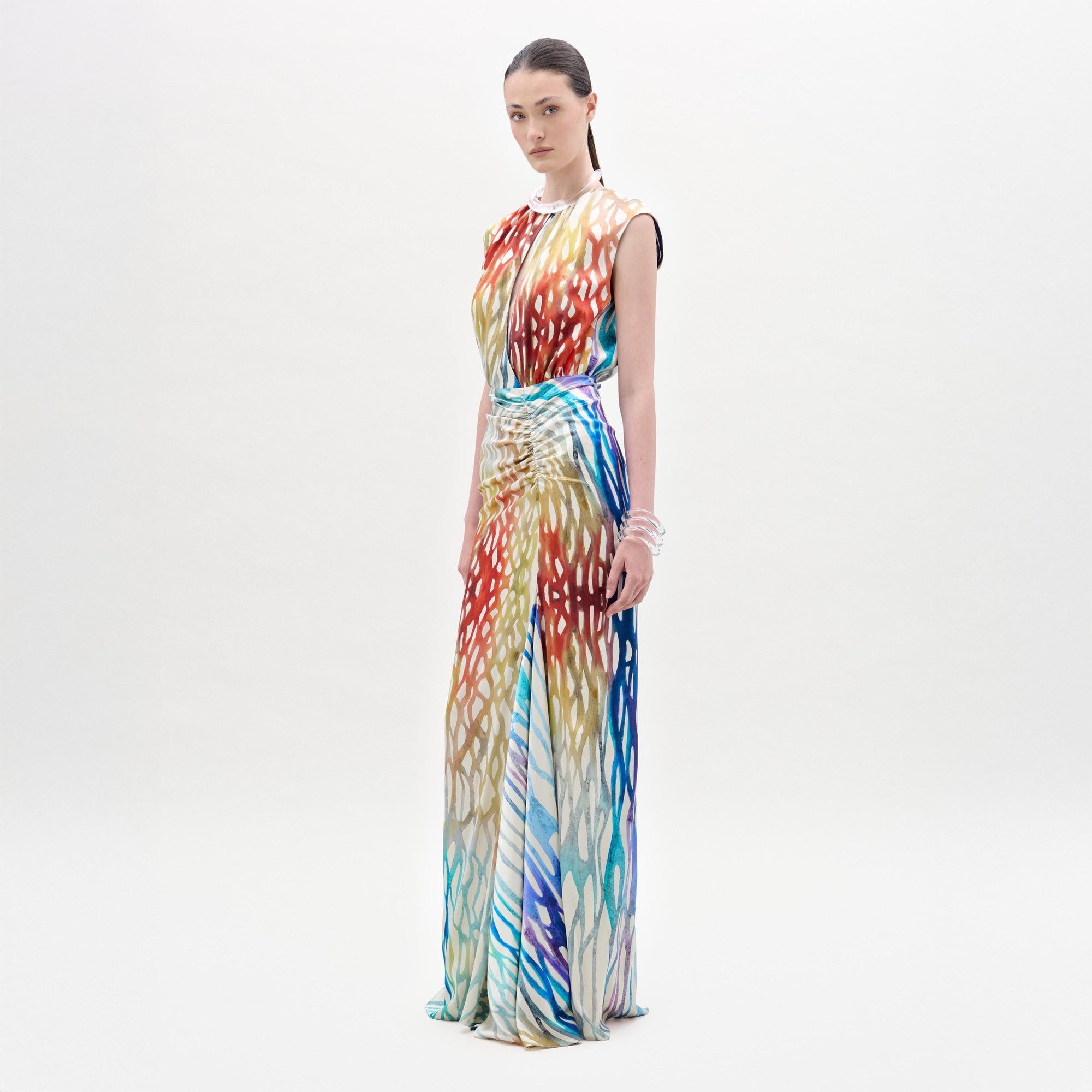 Una persona sobre un fondo blanco liso luce el vestido Samaira, un maxi vestido con escote de ojo de cerradura y vibrante estampado abstracto en rojo, amarillo, azul y blanco. Fabricado en Colombia. La persona lleva el pelo oscuro recogido y mira hacia un lado.