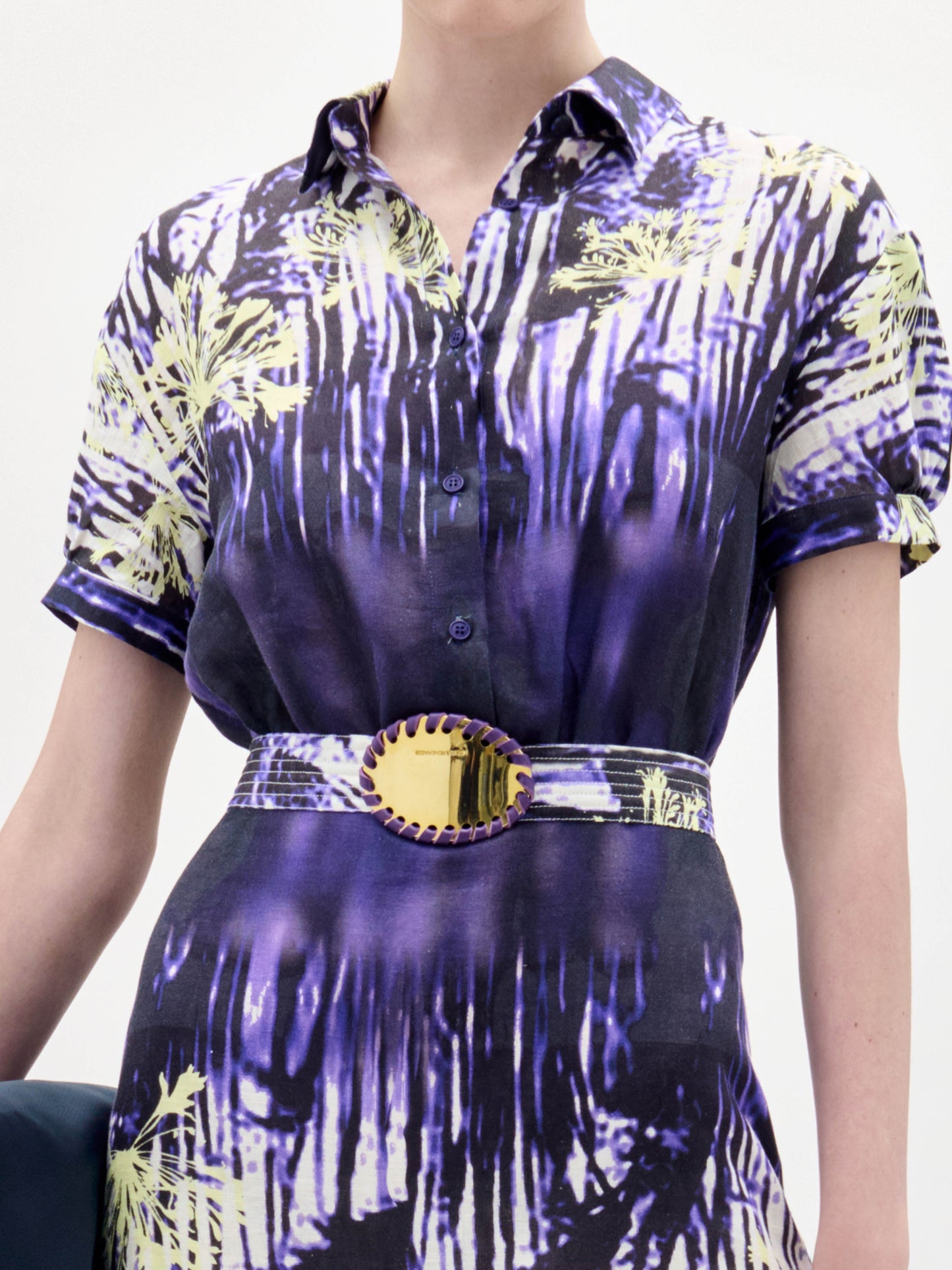 Una persona lleva el Vestido Lee, una prenda abotonada de manga corta con un estampado abstracto morado, amarillo y negro, ceñida a la cintura por un cinturón ancho con una gran hebilla circular.