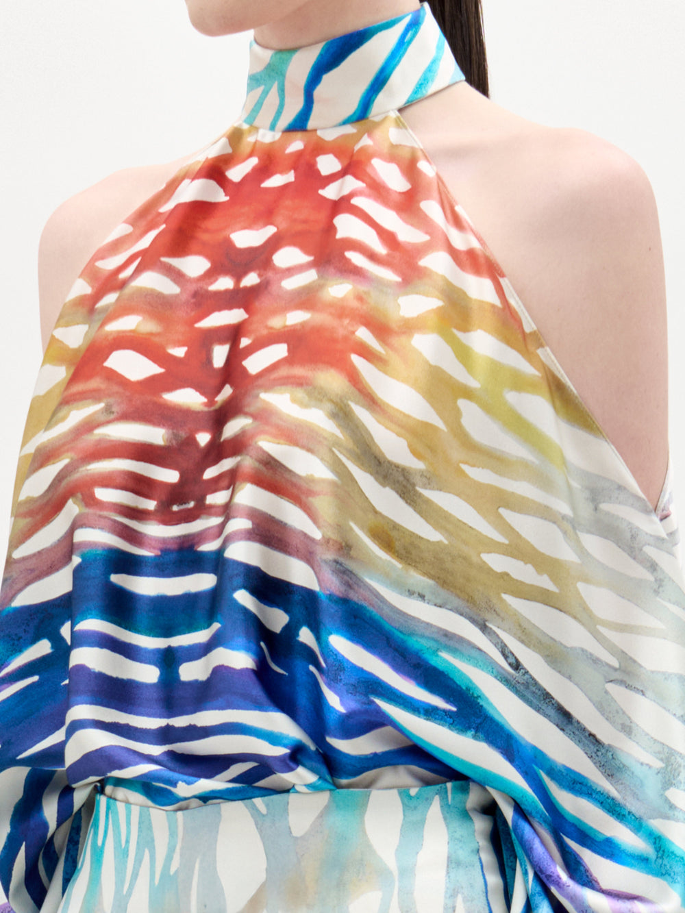 Una persona lleva la blusa Safira, un top sin mangas de cuello alto con un estampado abstracto de rayas rojas, amarillas, azules y blancas. La foto está recortada por el cuello y la parte superior de los brazos.