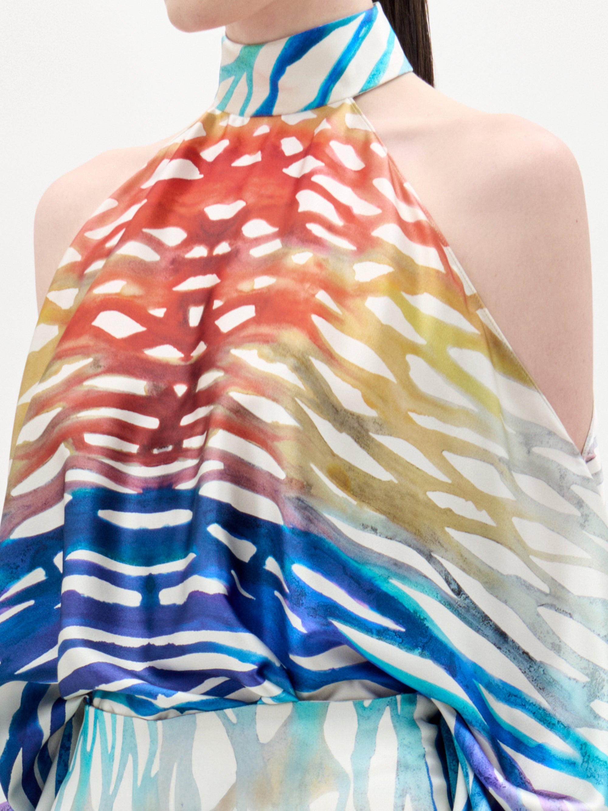 Una persona lleva la blusa Safira, un top sin mangas de cuello alto con un estampado abstracto de rayas rojas, amarillas, azules y blancas. La foto está recortada por el cuello y la parte superior de los brazos.