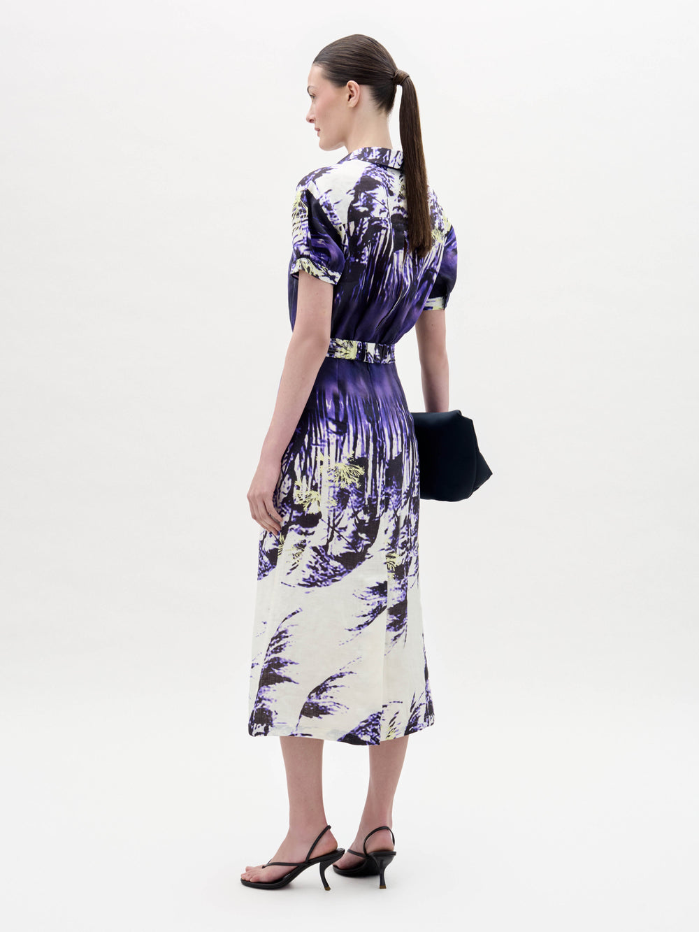 Una mujer aparece de perfil sobre un fondo blanco liso, con el vestido Lee -un vestido midi estampado en morado y blanco- combinado con sandalias de tacón negras y un bolso de mano oscuro. Lleva el pelo recogido en una coleta baja.