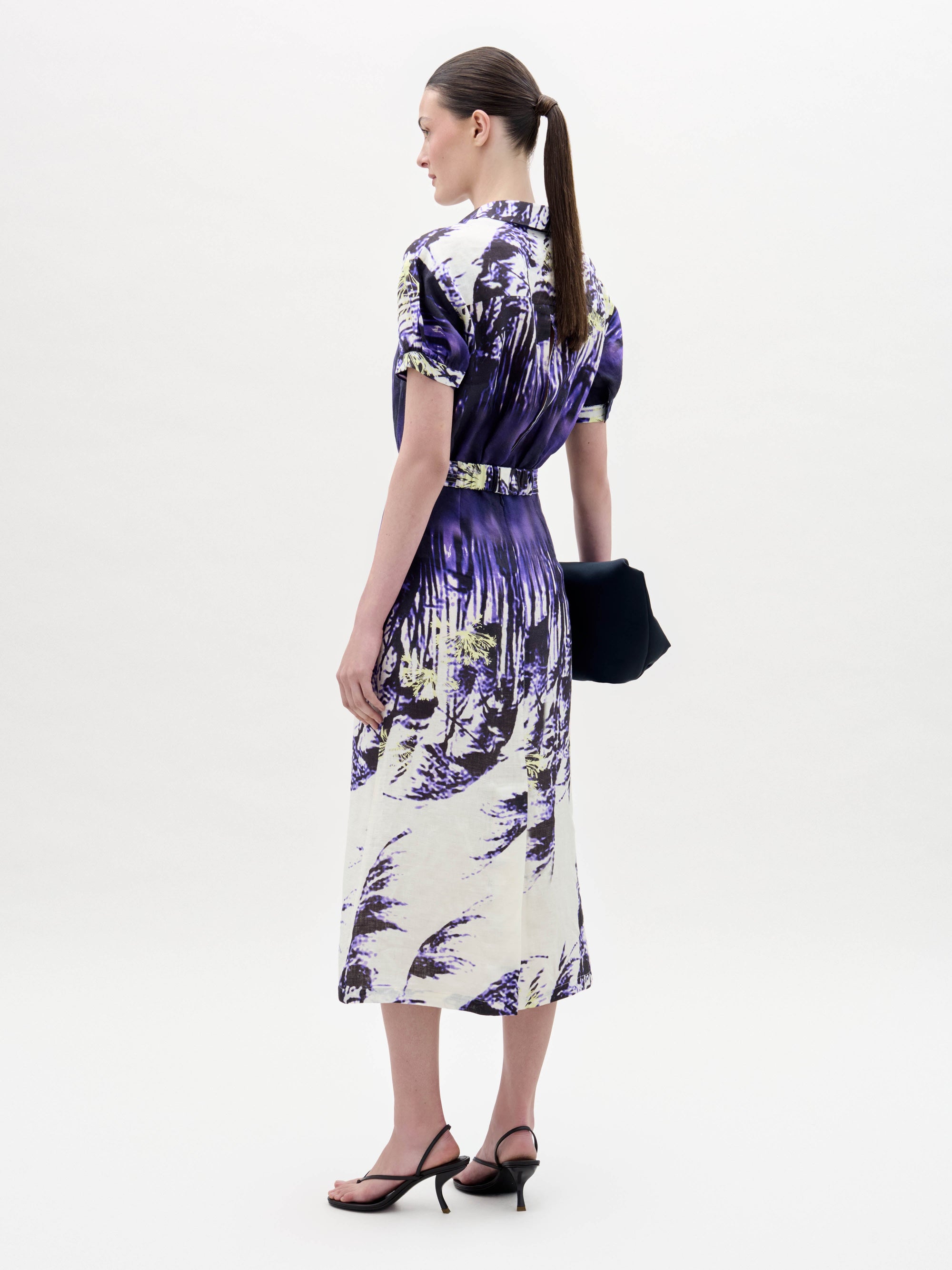 Una mujer aparece de perfil sobre un fondo blanco liso, con el vestido Lee -un vestido midi estampado en morado y blanco- combinado con sandalias de tacón negras y un bolso de mano oscuro. Lleva el pelo recogido en una coleta baja.