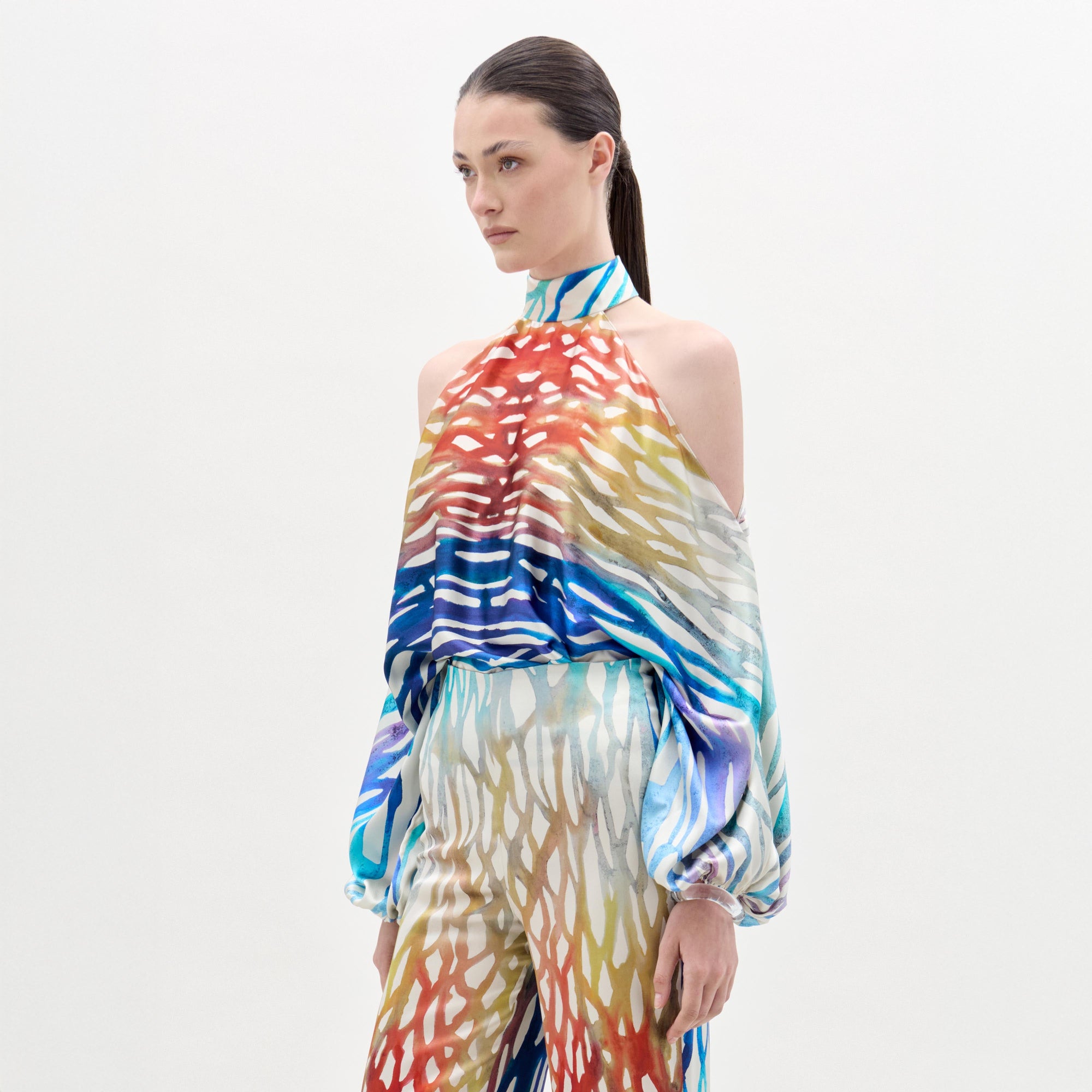 Una mujer con el pelo castaño liso recogido hacia atrás lleva la blusa Safira y unos pantalones anchos a juego, ambos en tejido italiano con estampado abstracto. Está de pie sobre un fondo blanco liso, mirando ligeramente a un lado.